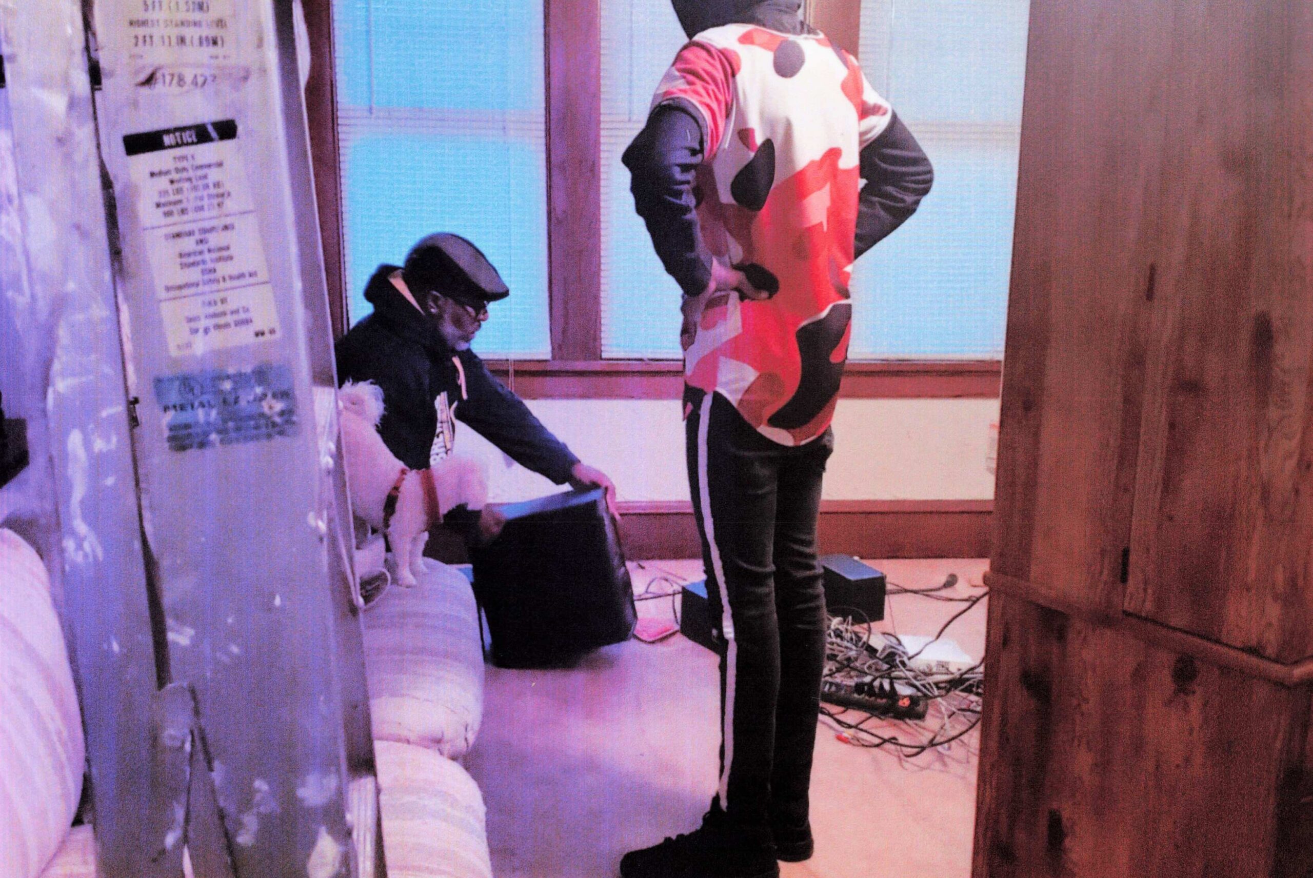 Two people in a cluttered room: one seated with a small white dog, the other standing in a red camouflage jacket; cables on the floor and a blue-tinted window in back.