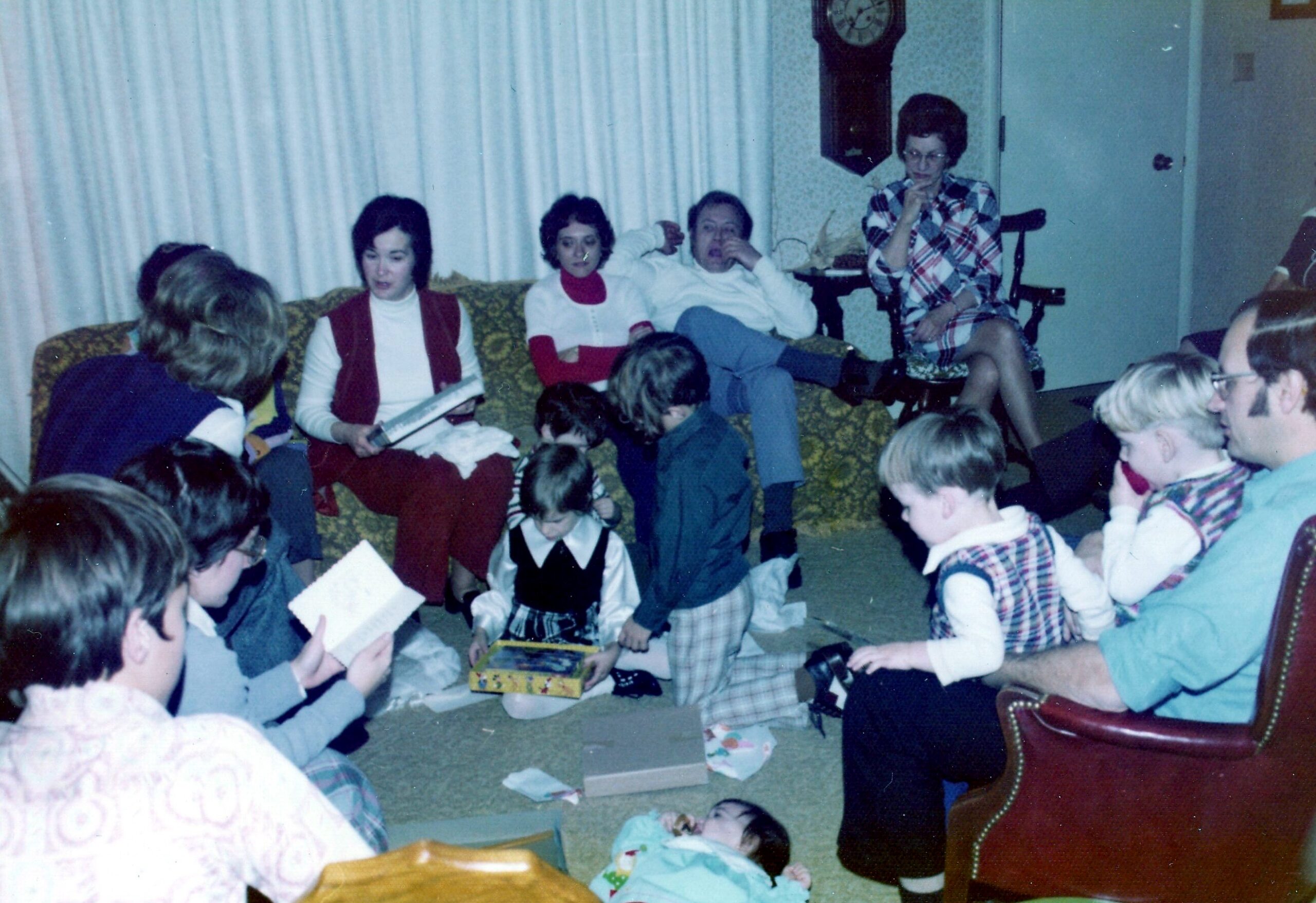 Kids sit on the floor and couch beside opened gifts at a family gathering; adults watch in a living room.