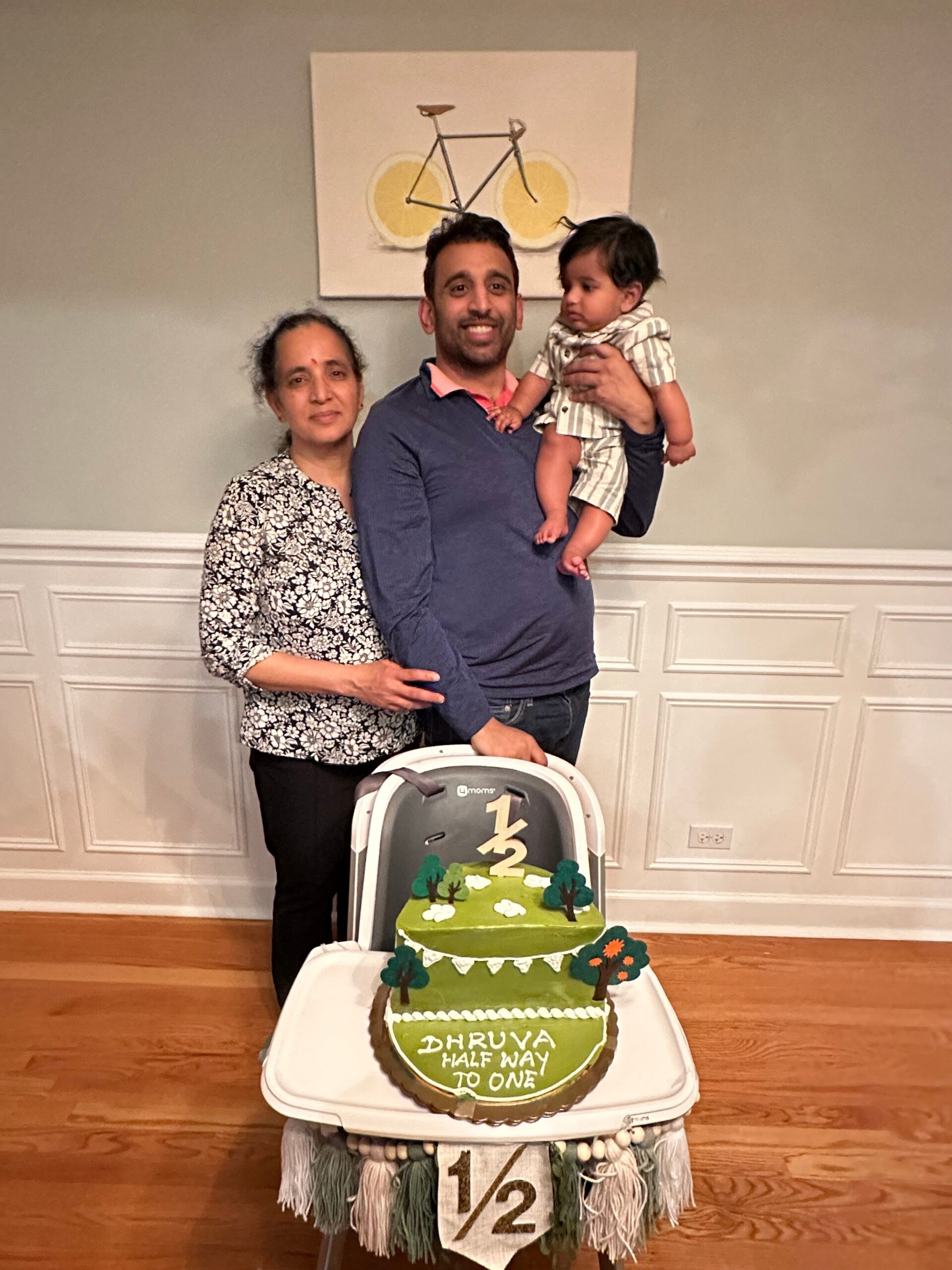 Family portrait with a man holding a baby and a grandmother beside him, in front of a half‑birthday cake that reads 'DHRUVA HALF WAY TO ONE' with a forest-themed topper on a high chair.