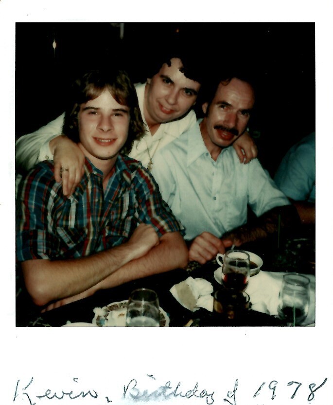 Three men sit closely at a table during a birthday celebration; one in a plaid shirt, others in light shirts, with drinks and plates in front.