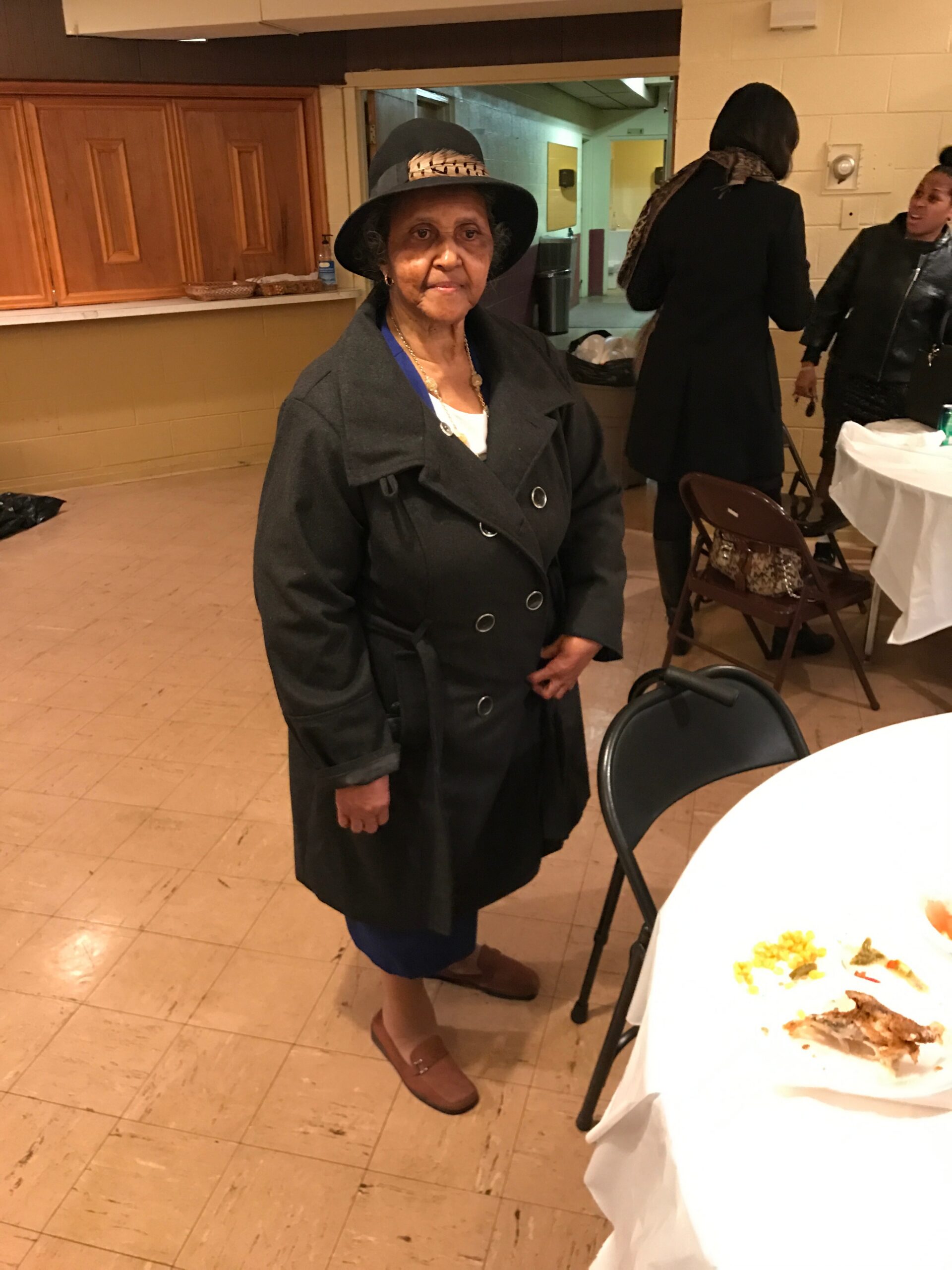 Older woman in a dark coat and wide-brim hat stands near a table with food at a community event.