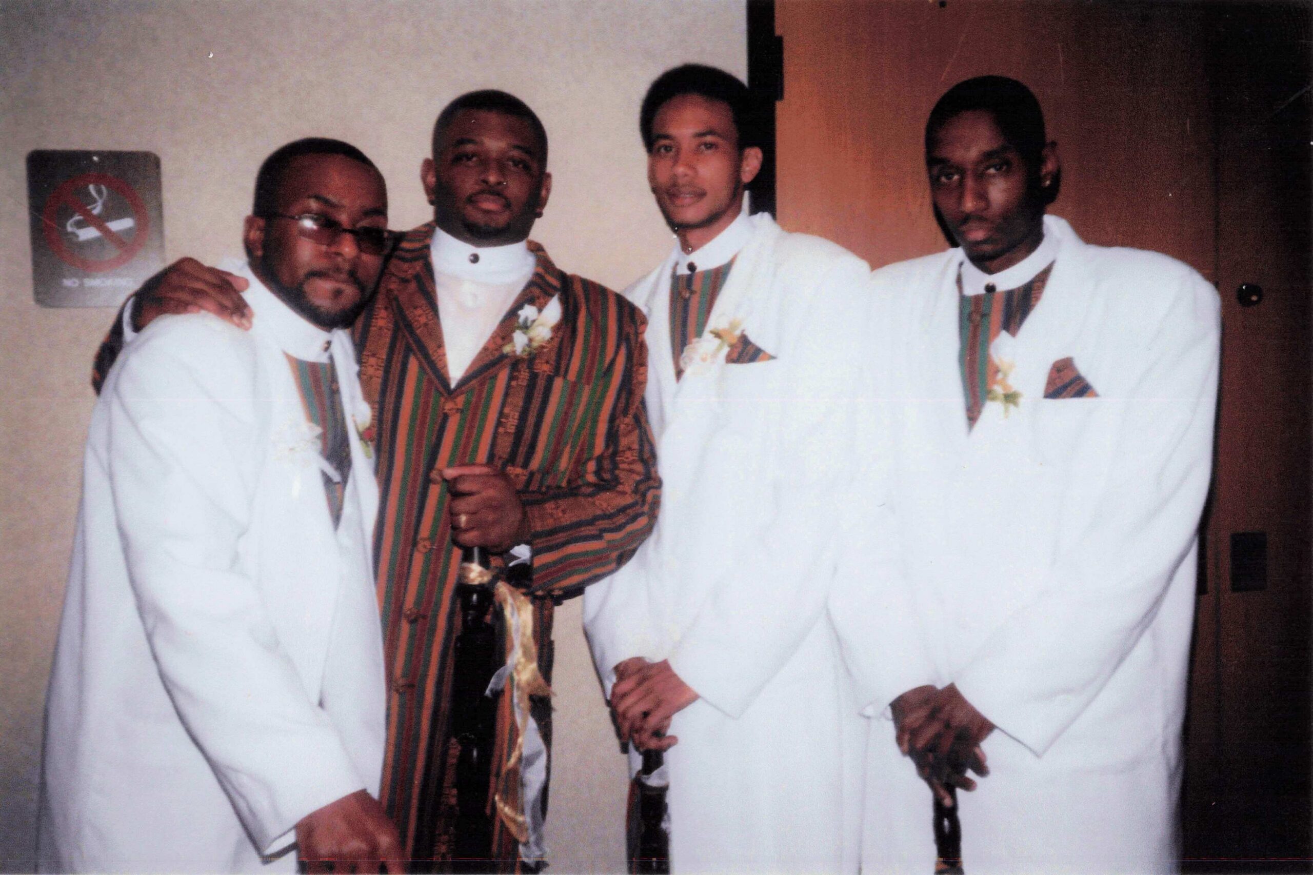 Group portrait of four men in formal attire posing together at what appears to be a ceremony; three wear white suits with boutonnières and one wears a brown striped suit with a ring and a sash-like belt.