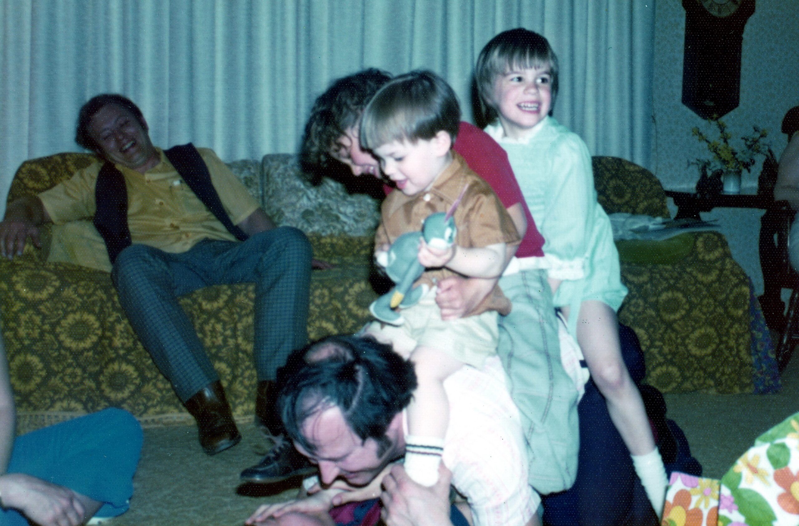 Children playing and laughing while adults relax on a patterned sofa in a dimly lit living room, with a plush toy visible in a child’s hands.