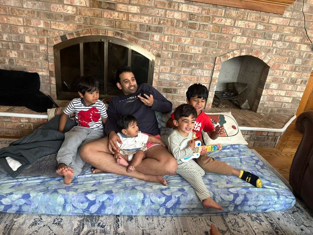 A man sits on a blue floral sofa with five young children in a living room with a brick fireplace.