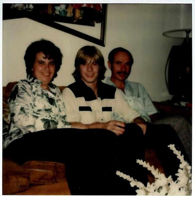 Three people (woman on left, blond man in middle, man with mustache on right) seated on a brown couch in a living room, smiling at the camera.