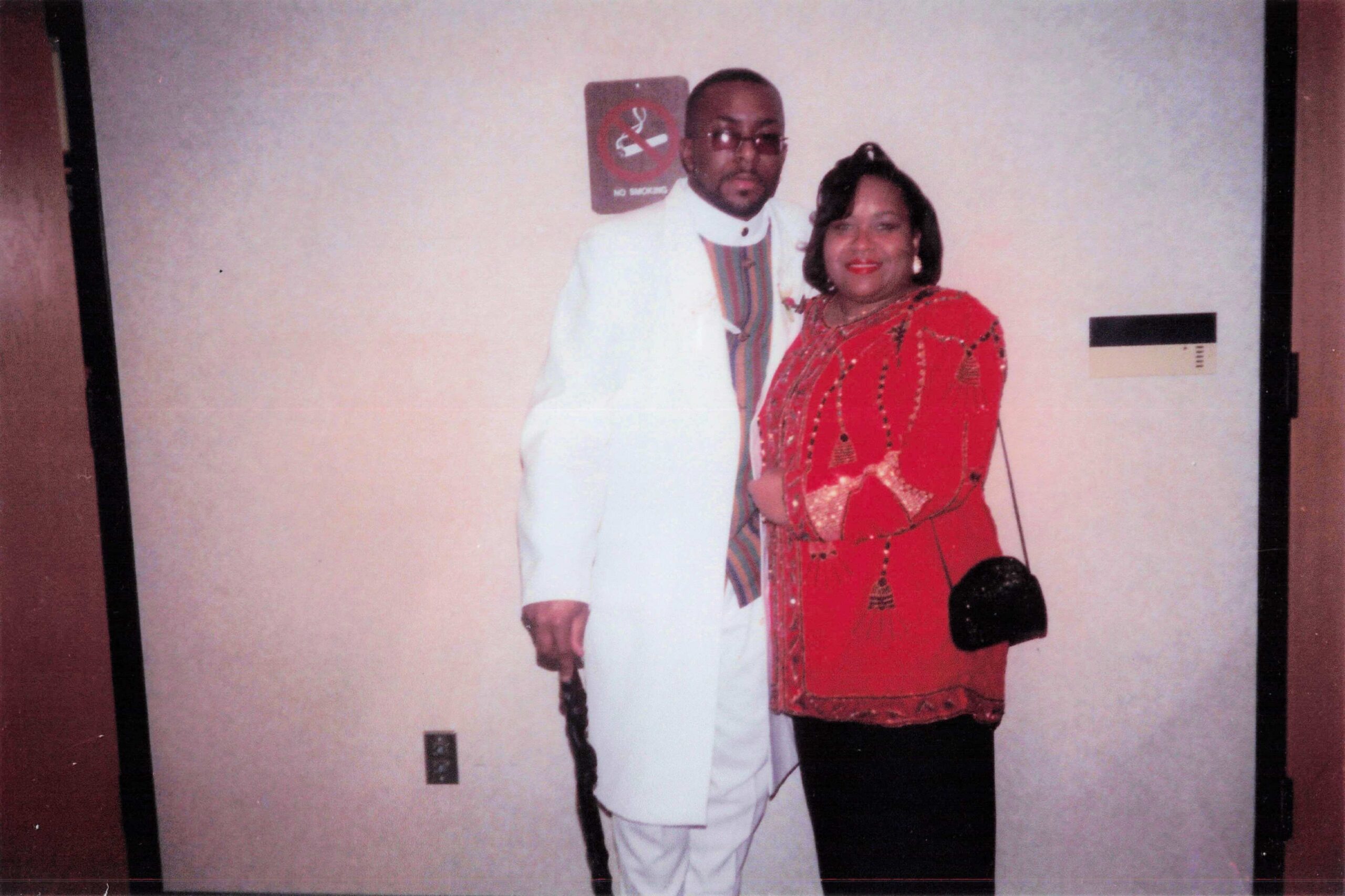 Couple posing for a photo indoors; man in a white suit with a colorful scarf and a woman in a red patterned outfit.