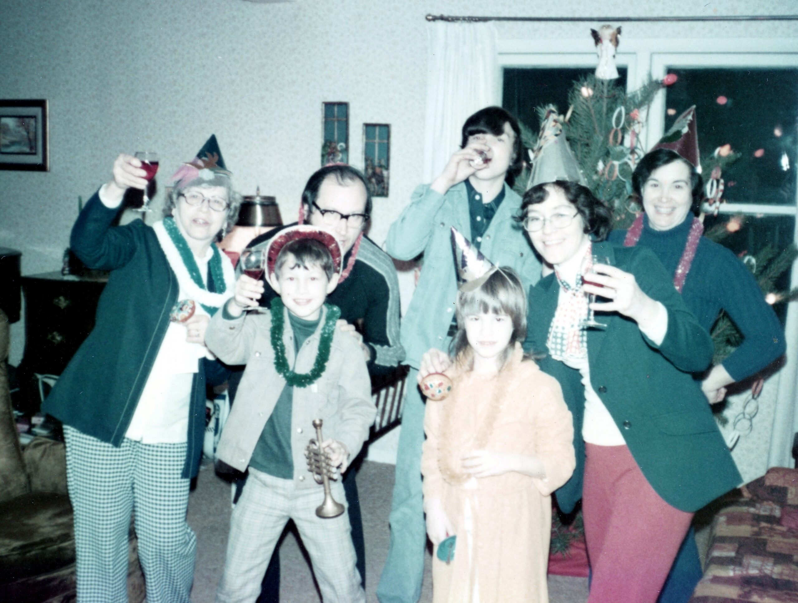 Group of people at a party raising glasses, wearing hats and beads near a Christmas tree in the background and festive decorations quietly visible behind them.