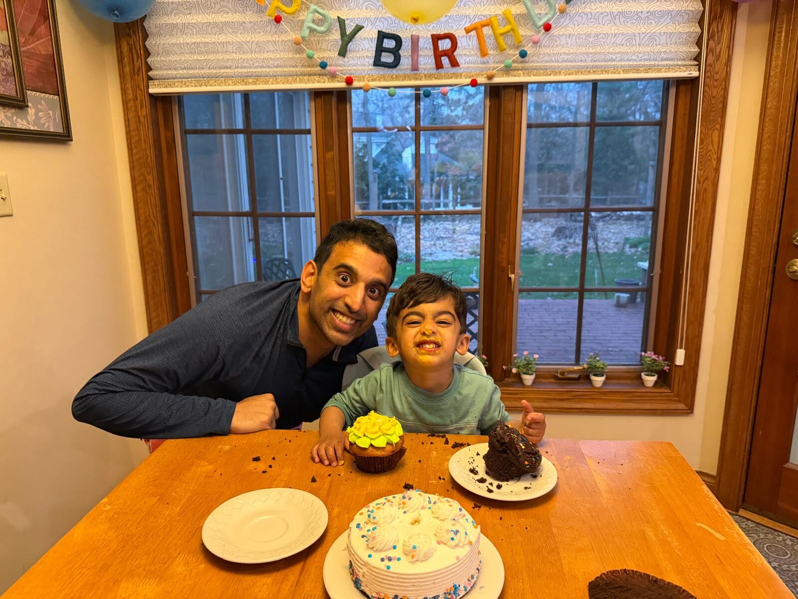 Two people smiling at a birthday table, a man leaning beside a child with cake frosting smeared on his face, a colorful birthday banner above them.