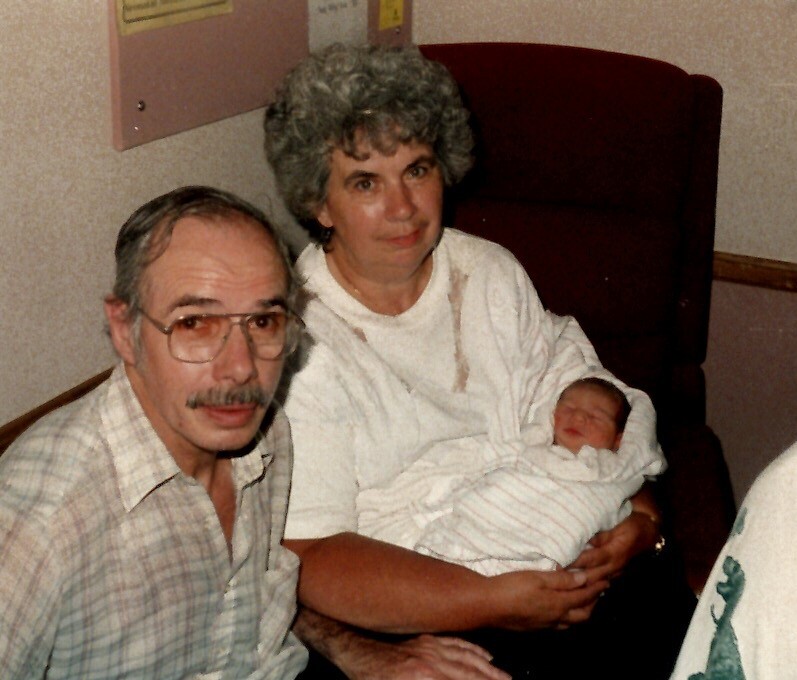 Older couple sitting in a hospital room, holding a newborn baby wrapped in a white blanket.