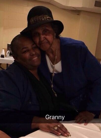 Granny wearing a hat stands beside a younger woman at a table, both smiling for the photo.