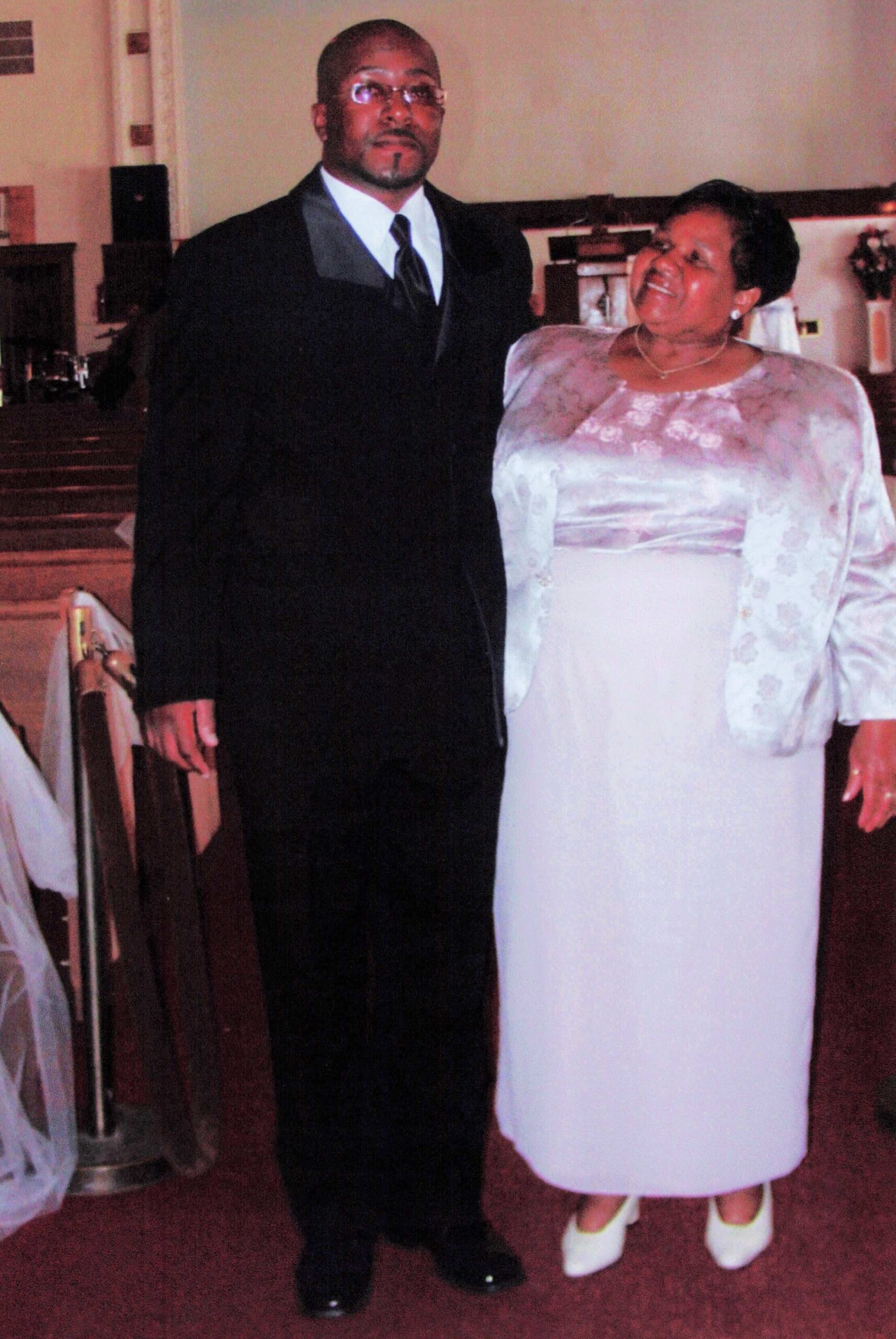 Couple in formal wear posing together inside a church, the man in a black suit and the woman in a lavender dress smile at the camera.