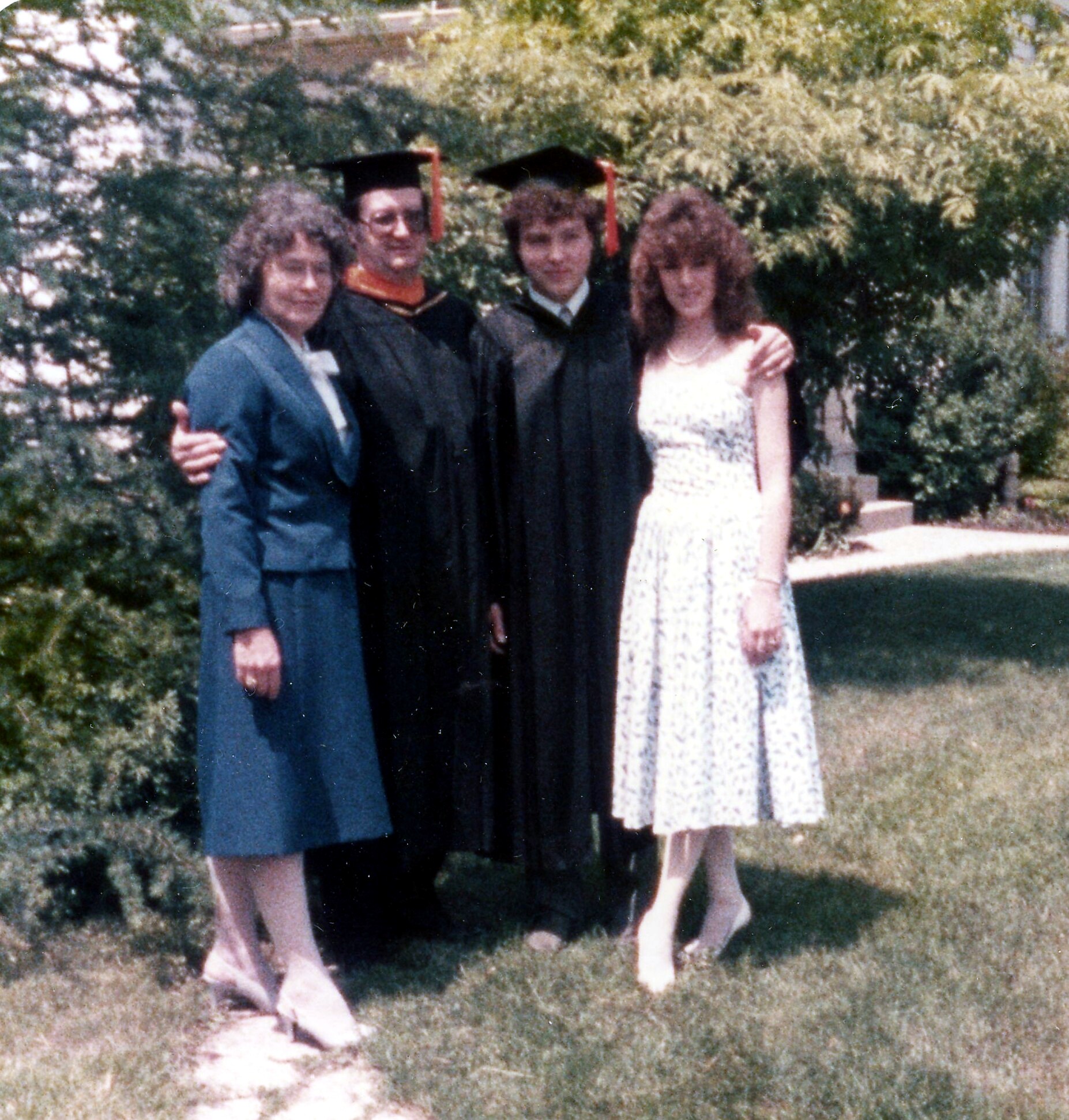 Four people posing outdoors for a graduation photo: two graduates in black gowns and mortarboards with red tassels, flanked by two others in formal attire against a leafy background.