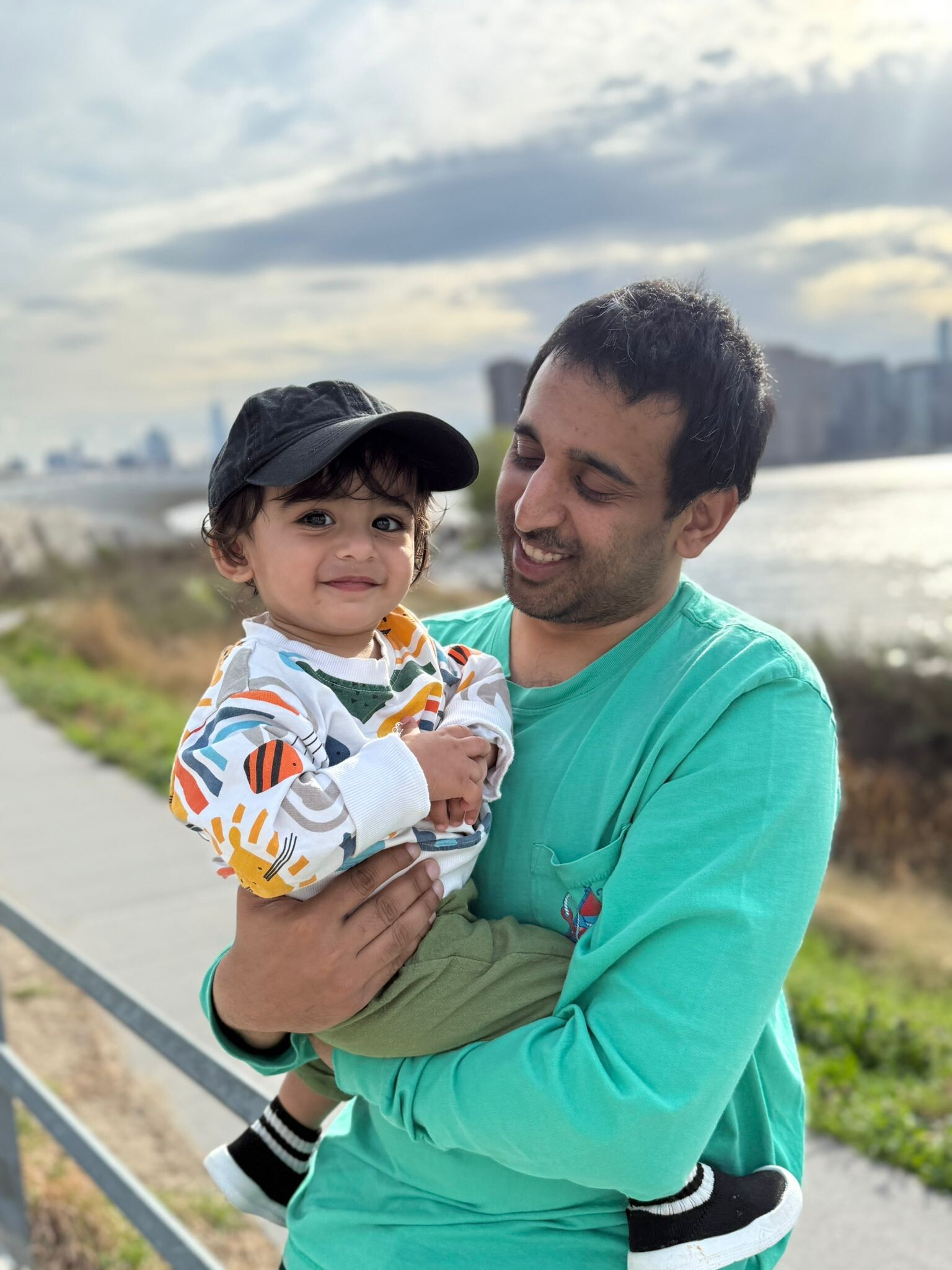 Man in a turquoise shirt holding a smiling toddler in a colorful sweater, with a waterfront and distant city skyline in the background.
