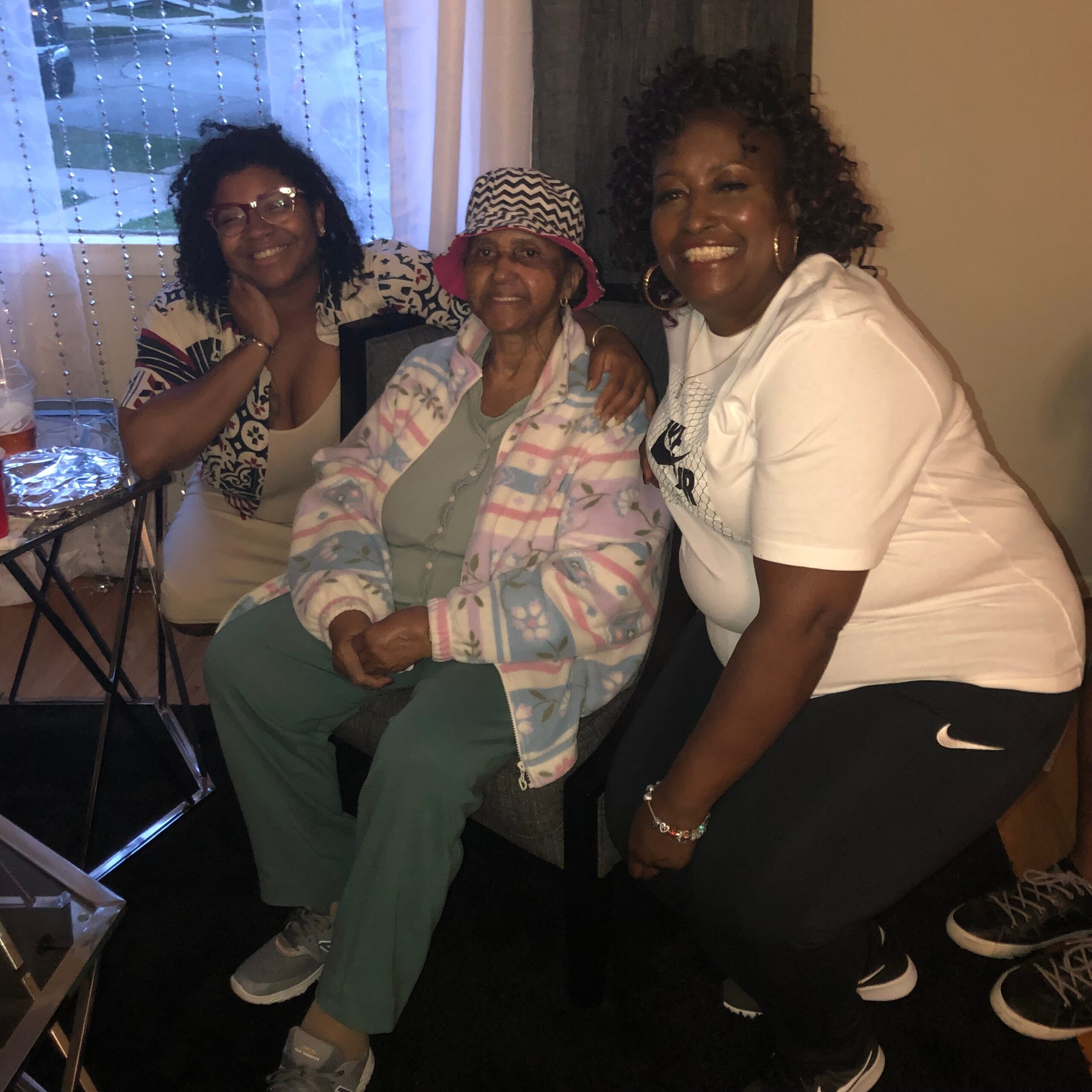 Three smiling women posing together on a couch indoors, with the center woman wearing a hat and colorful cardigan.