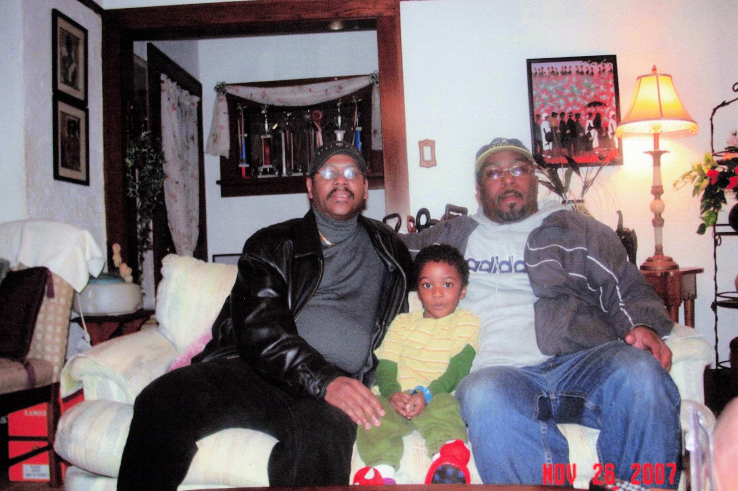 Family portrait of two men and a child sitting on a sofa in a cozy living room, with a lamp and wall art in the background.