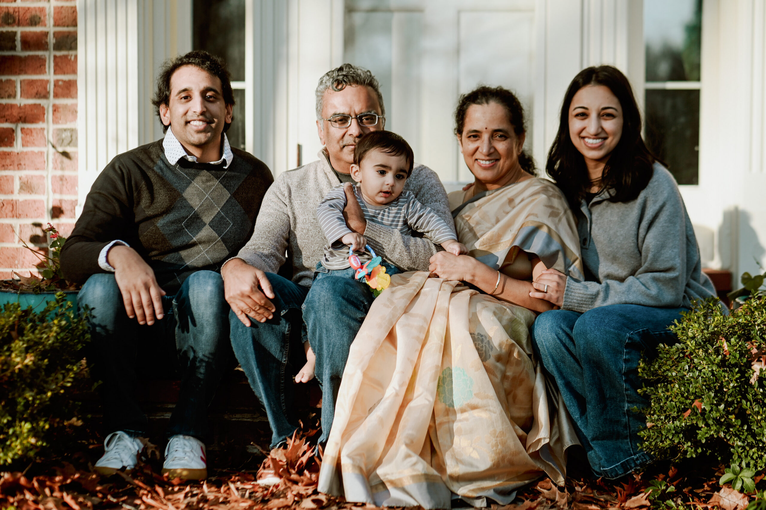 Five-person family gathered on front porch steps, smiling; baby sits on grandfather's lap with autumn leaves on the ground nearby.