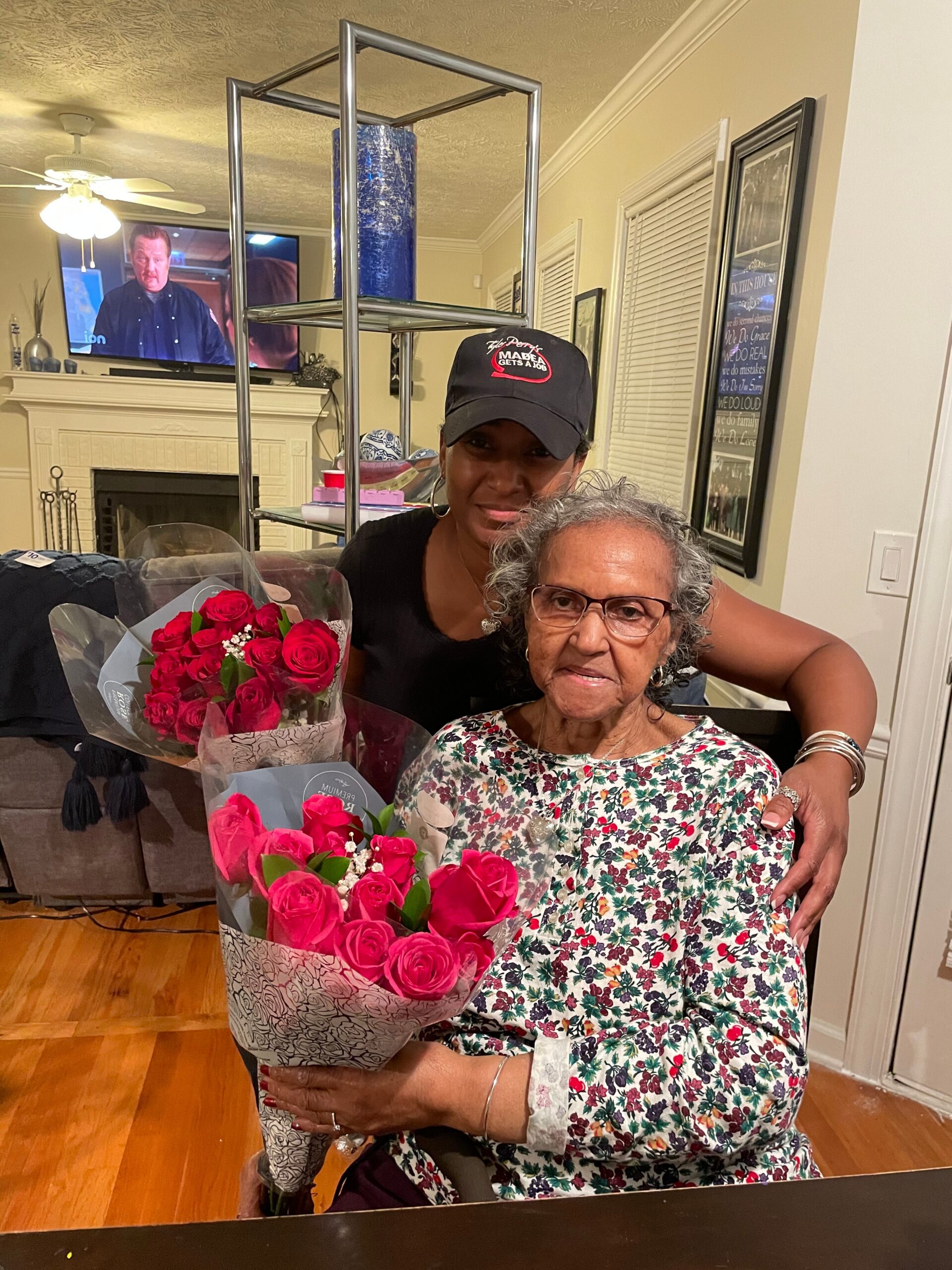 Two women pose indoors: an elderly woman in a floral dress sits, while a younger woman in a black cap hugs her, holding a bouquet of red roses in front.