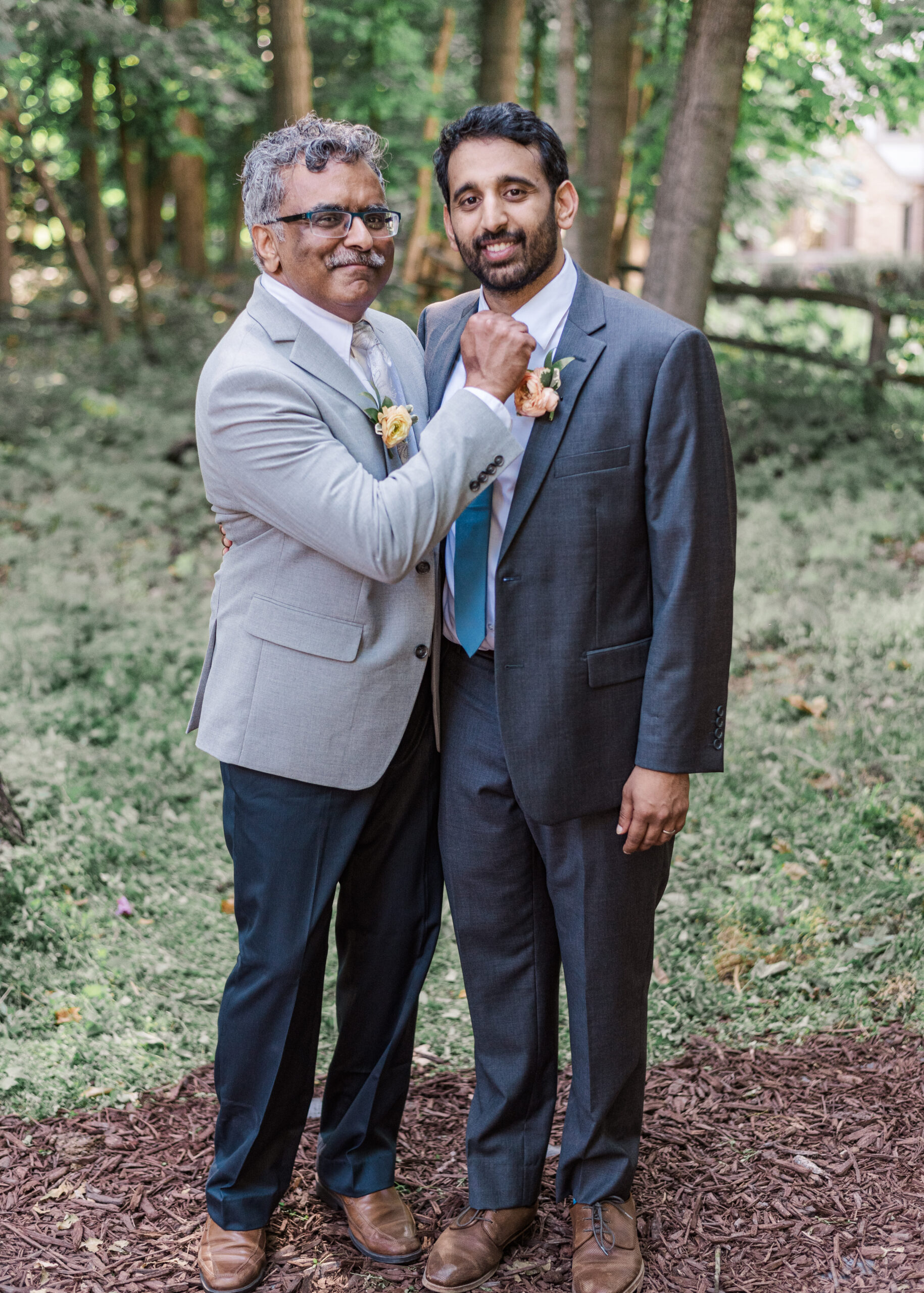 Two men in suits stand together outdoors in a wooded area; the older man adjusts the younger man's tie and smiles at the camera.