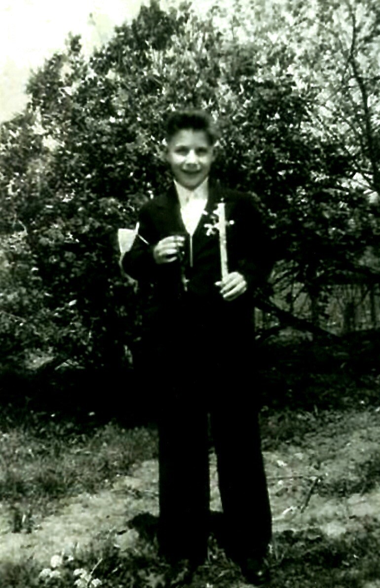 Boy in a dark suit outdoors, smiling, holding a single flower in one hand.