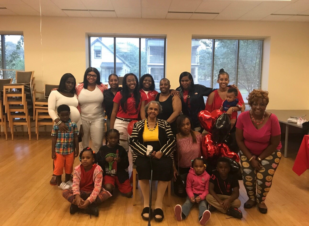 Group of diverse women and children pose together for a community celebration in a bright room, with an elderly woman seated center and red balloons nearby.