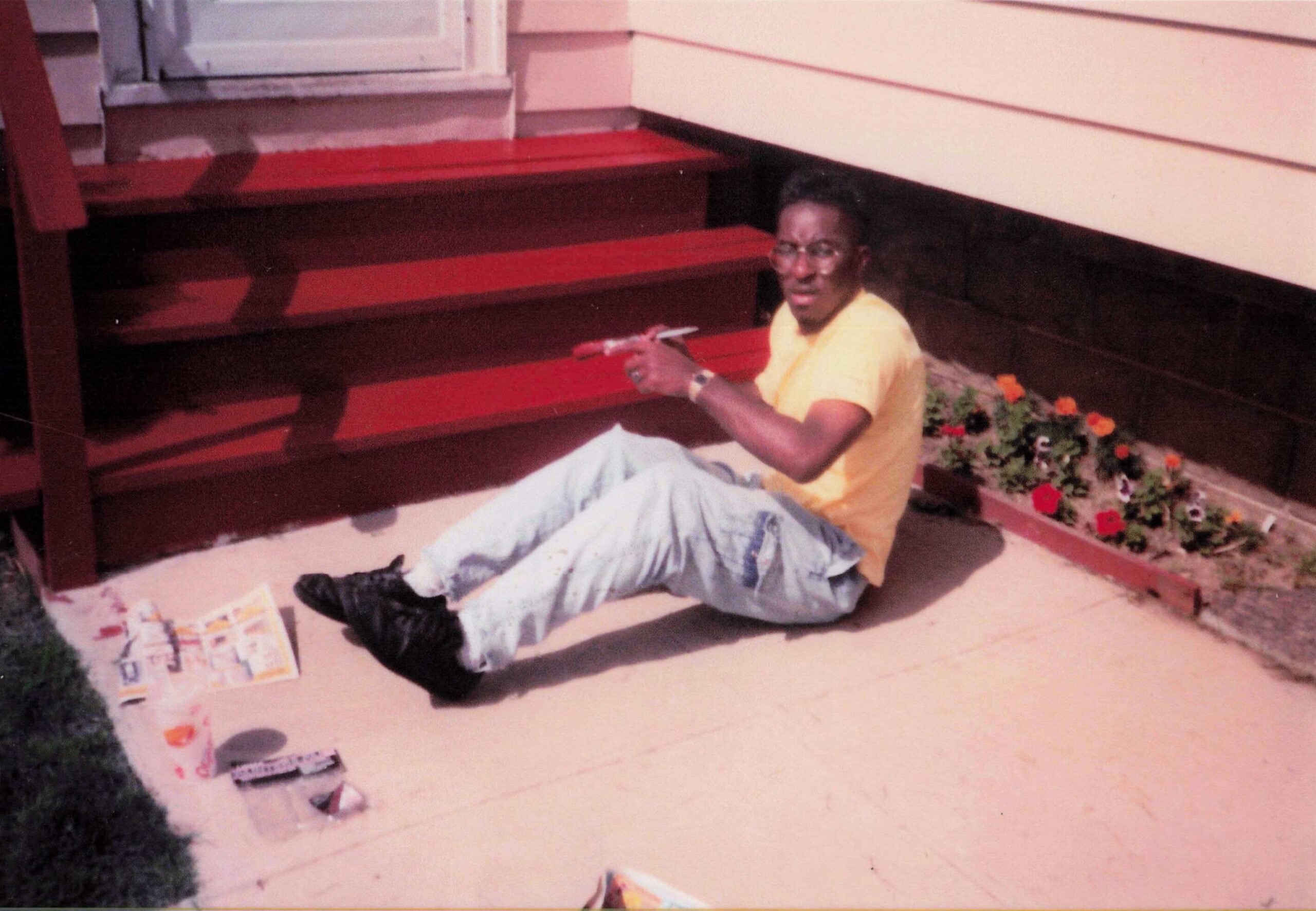 Man in a yellow shirt sitting on red wooden steps outside a house, pointing at the camera, wearing sunglasses.