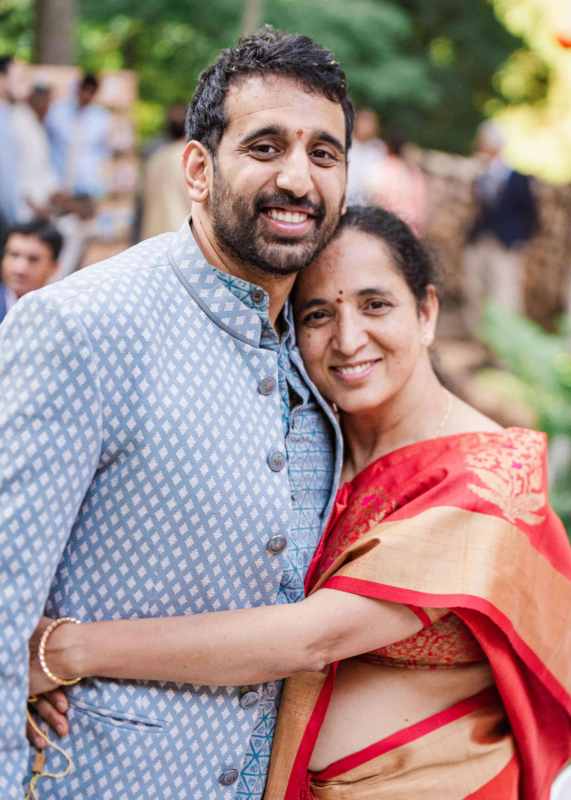 A smiling couple embraces outdoors, with the man in a light blue patterned kurta and the woman in a red saree.