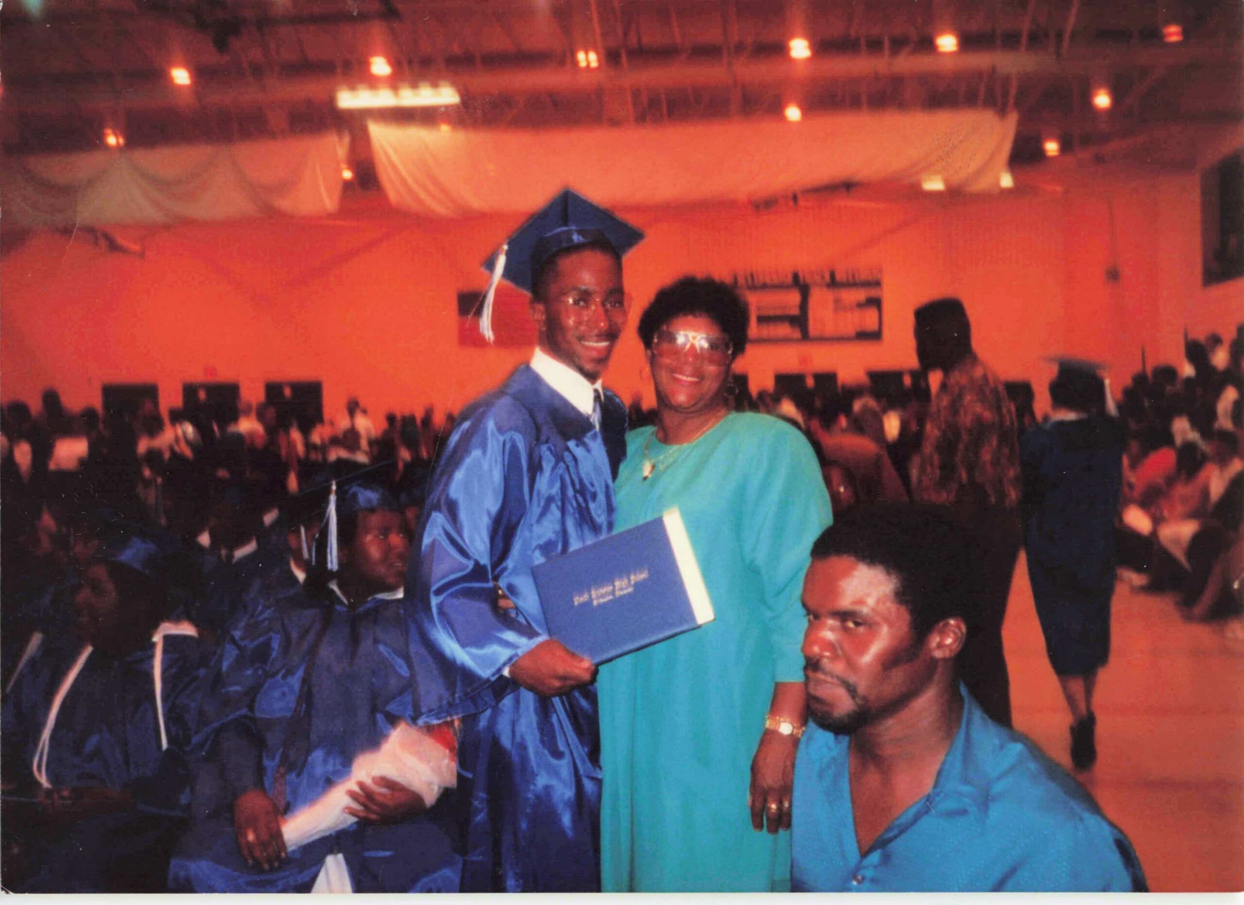 Graduate in blue gown with cap posing with a smiling woman in a green shirt at a crowded ceremony hall orange lighting scene.