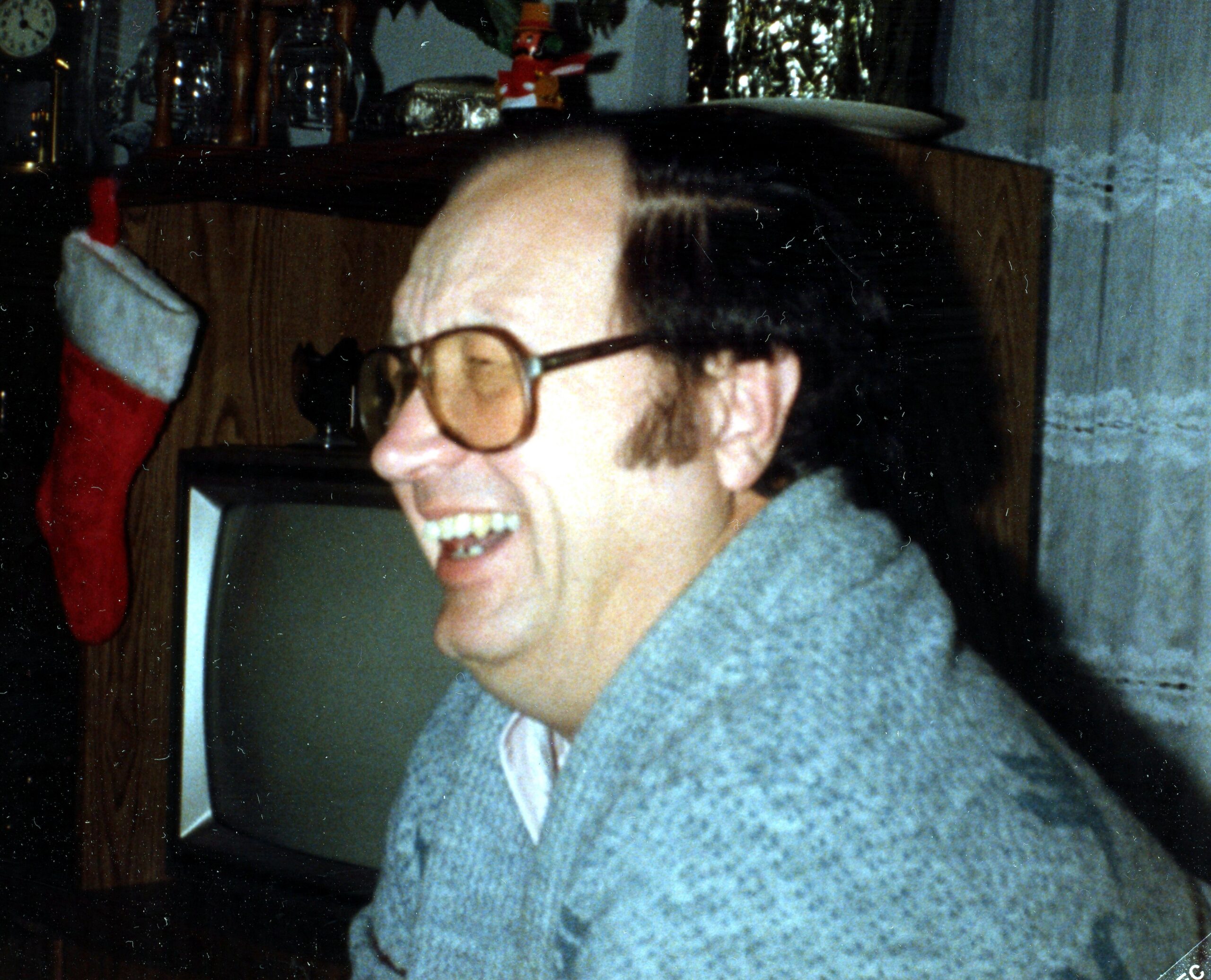 Smiling man wearing glasses in a cozy living room with a Christmas stocking hanging on the left side of a wooden cabinet behind him.