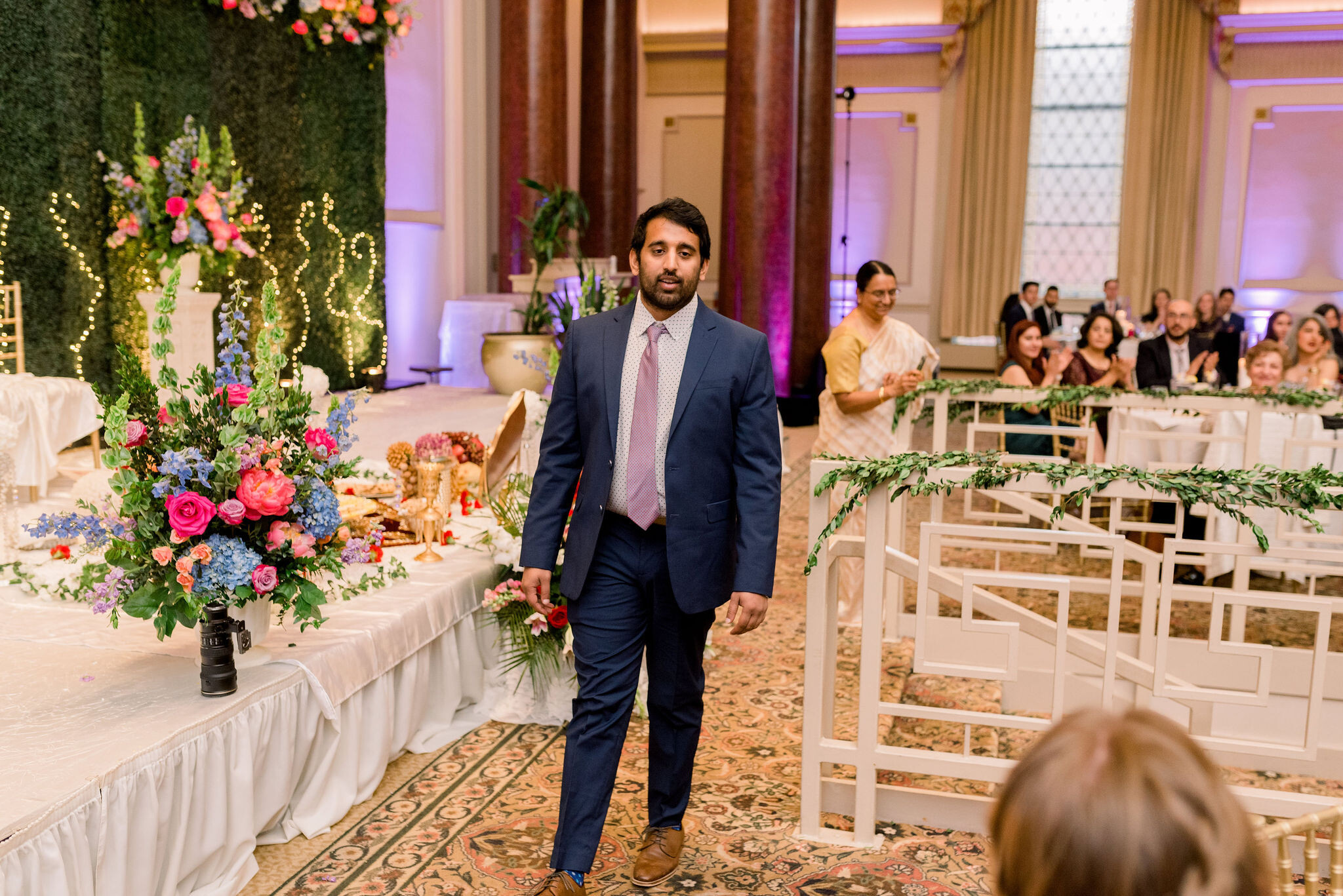 Man in a navy suit walks down an aisle at a wedding reception as guests sit at tables in the background with floral decor.