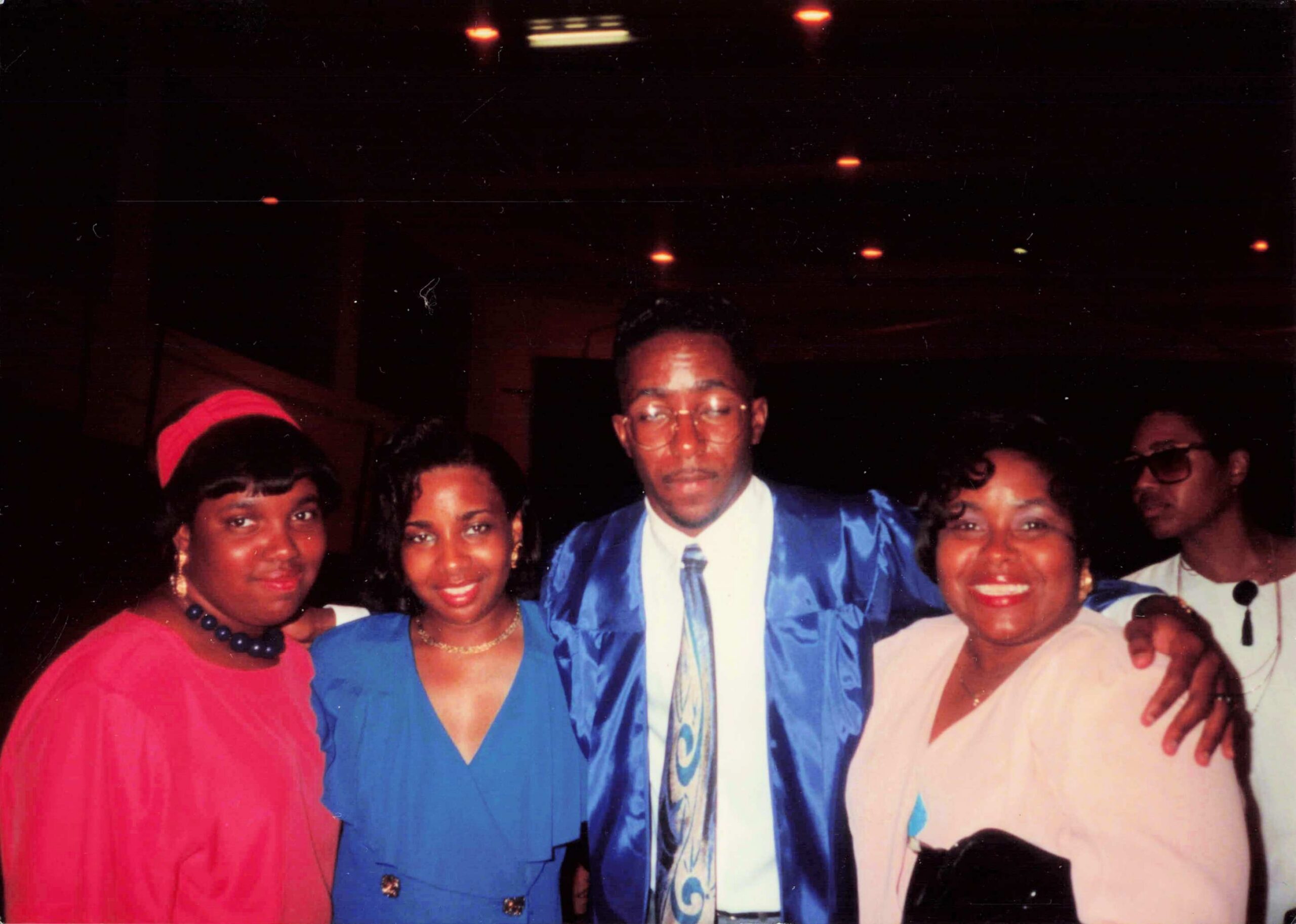 Group of friends at a party posing for a photo; a man in a blue suit stands center with others around him, all smiling.