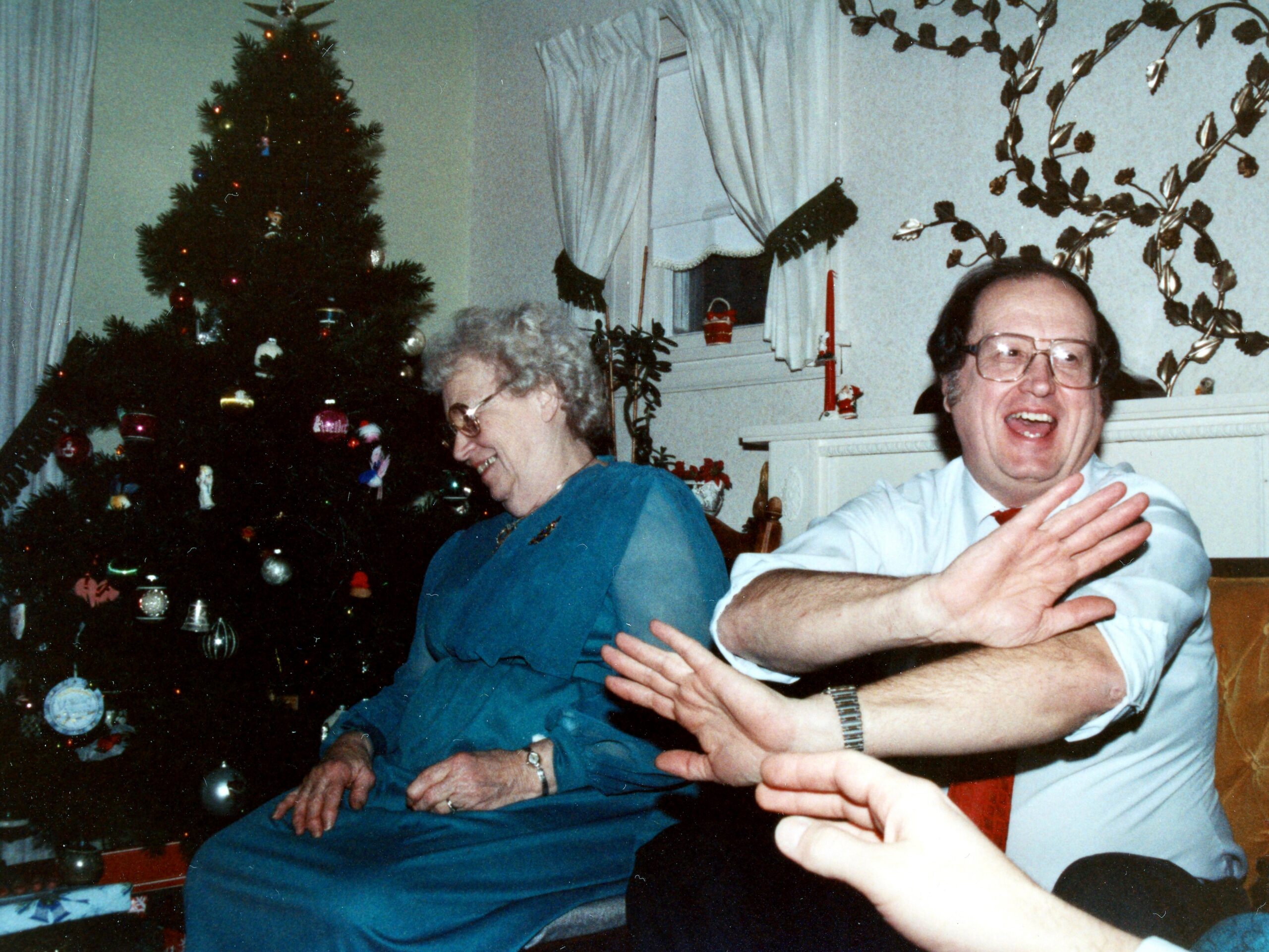 An elderly couple seated by a Christmas tree, smiling and laughing indoors as hands reach toward the camera.