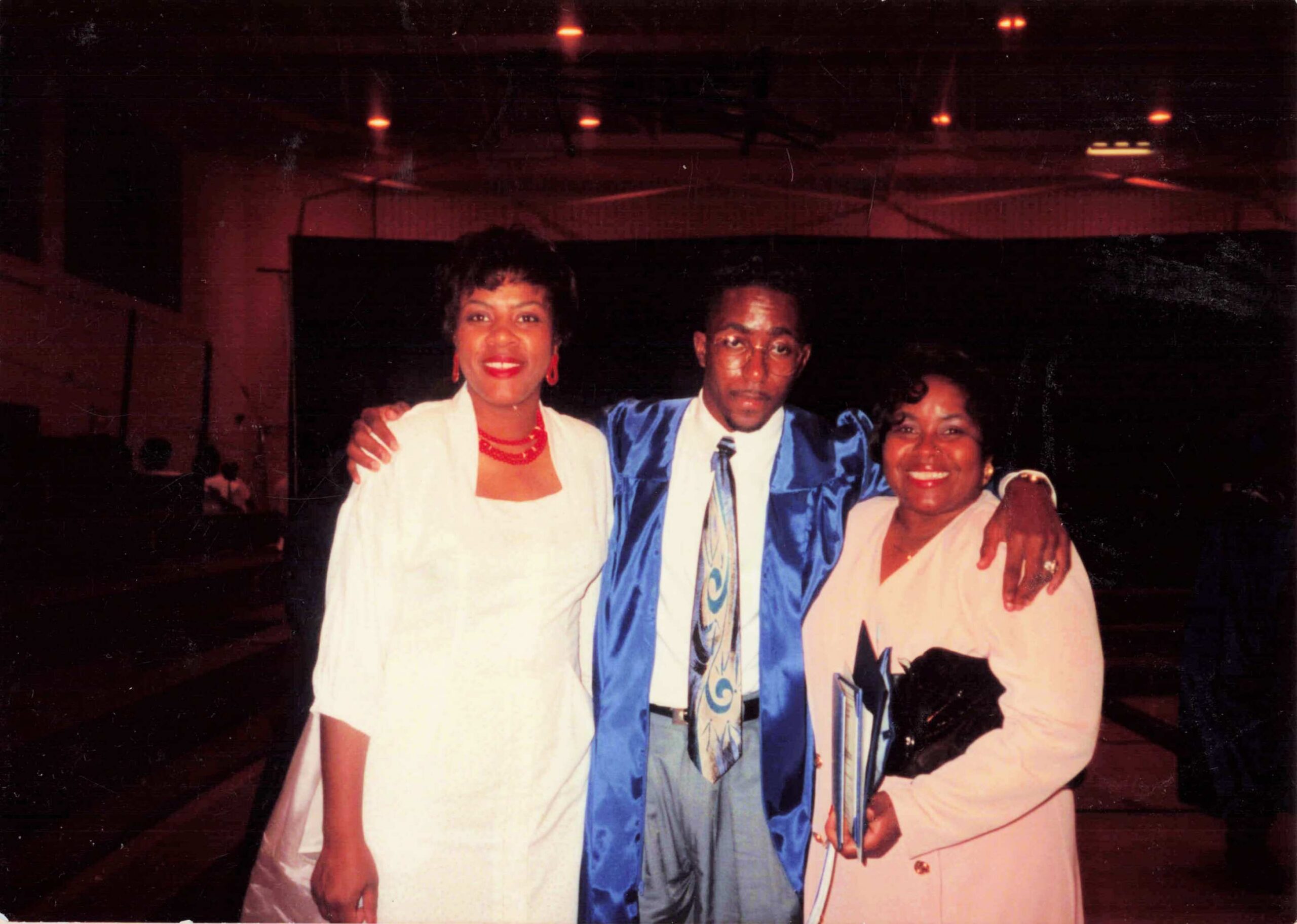Three people pose together at a formal event; a man in a blue satin suit stands between two women in white outfits, all smiling.