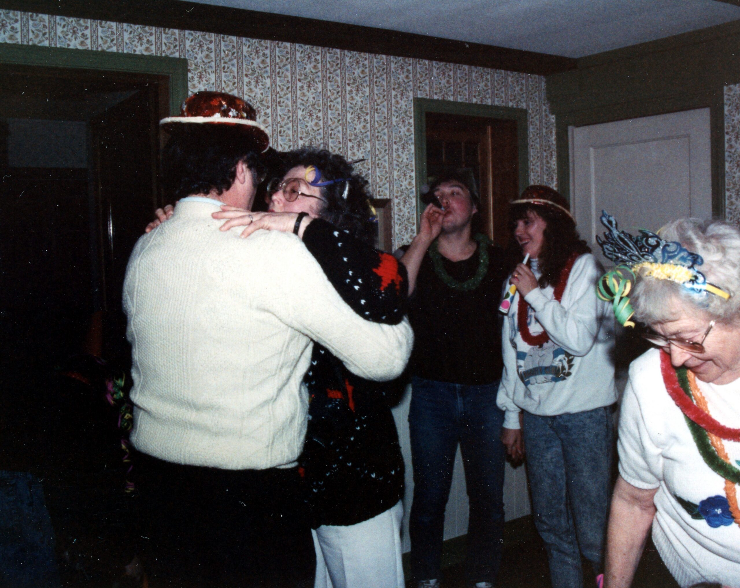 People dancing at a home party indoors, wearing festive hats and colorful costumes against floral wallpapered walls, with several guests socializing.