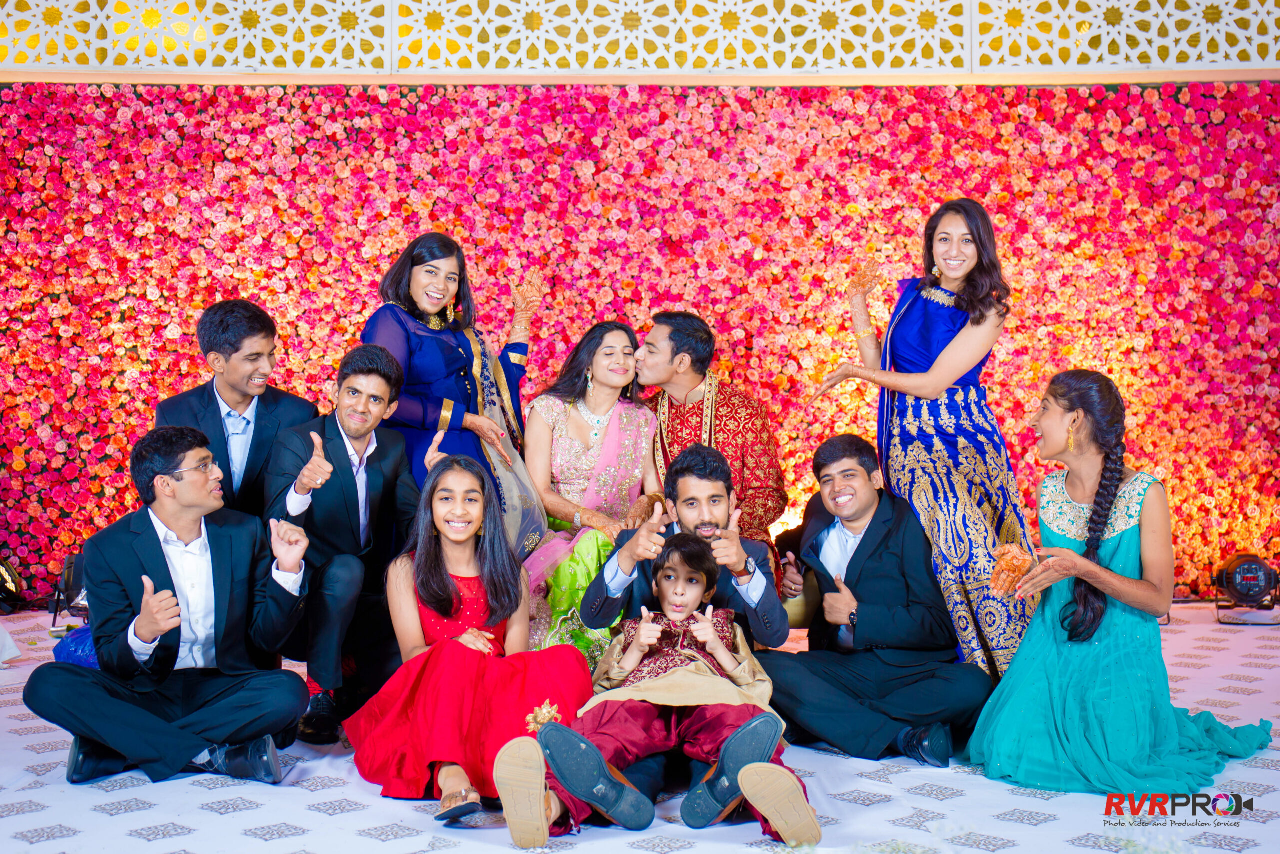 Group of friends and family in colorful Indian outfits posing and smiling in front of a pink flower wall at a celebration studio backdrop.