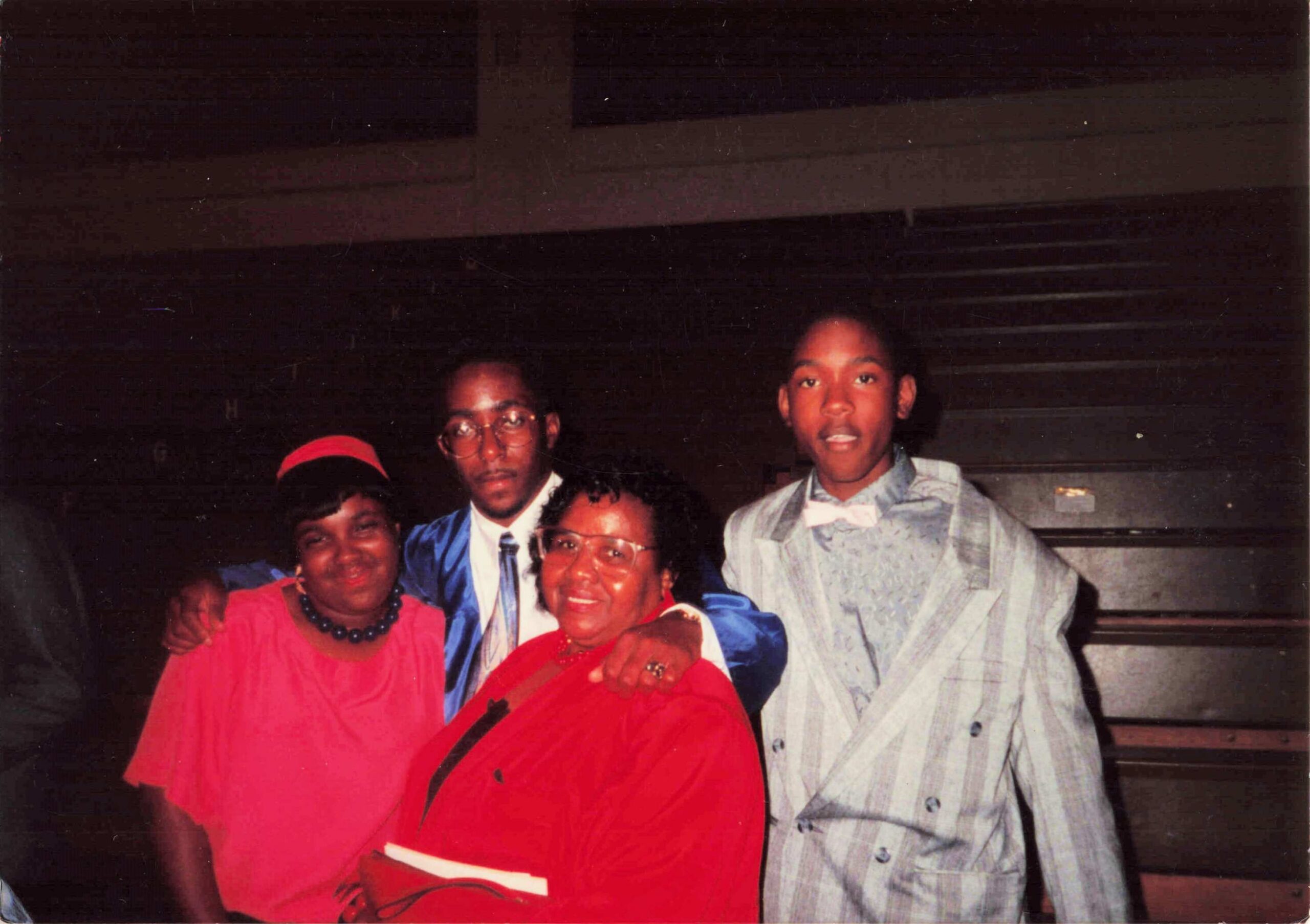 Group of friends posing together at a social gathering, smiling and dressed in bright red outfits and suits in a dim indoor setting.