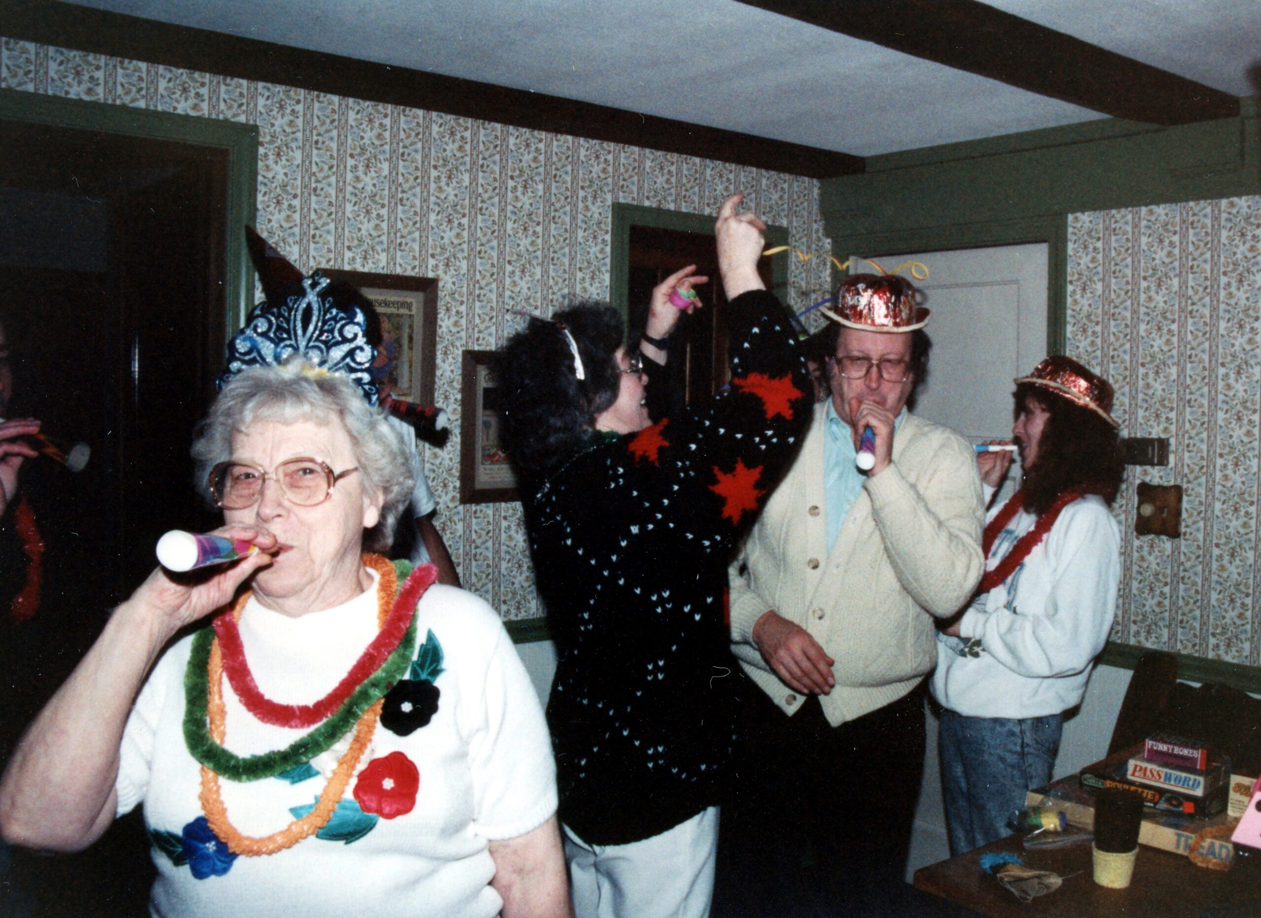 Older woman in foreground blowing a party horn, wearing colorful leis at a festive indoor party with people wearing hats in the background.