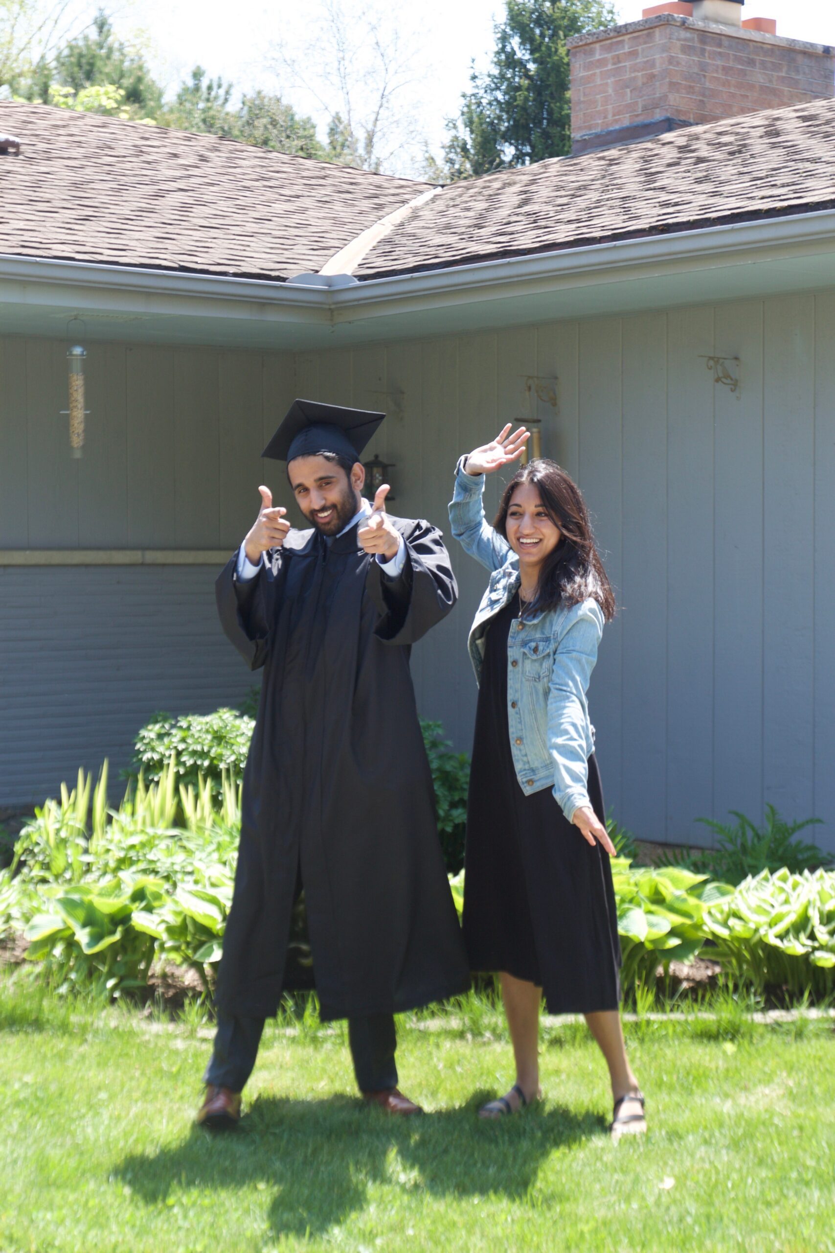 Two graduates celebrate outdoors: a man in cap and gown pointing at the camera while a woman in a denim jacket waves beside him in a garden setting.