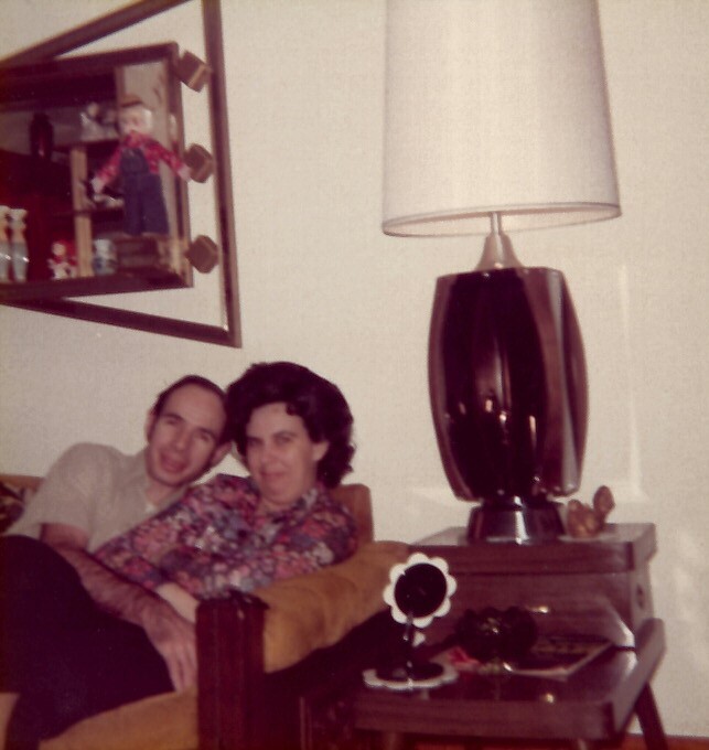 Man and woman sitting close on a mustard sofa in a retro living room, with a large black lamp on a side table and mirror above.