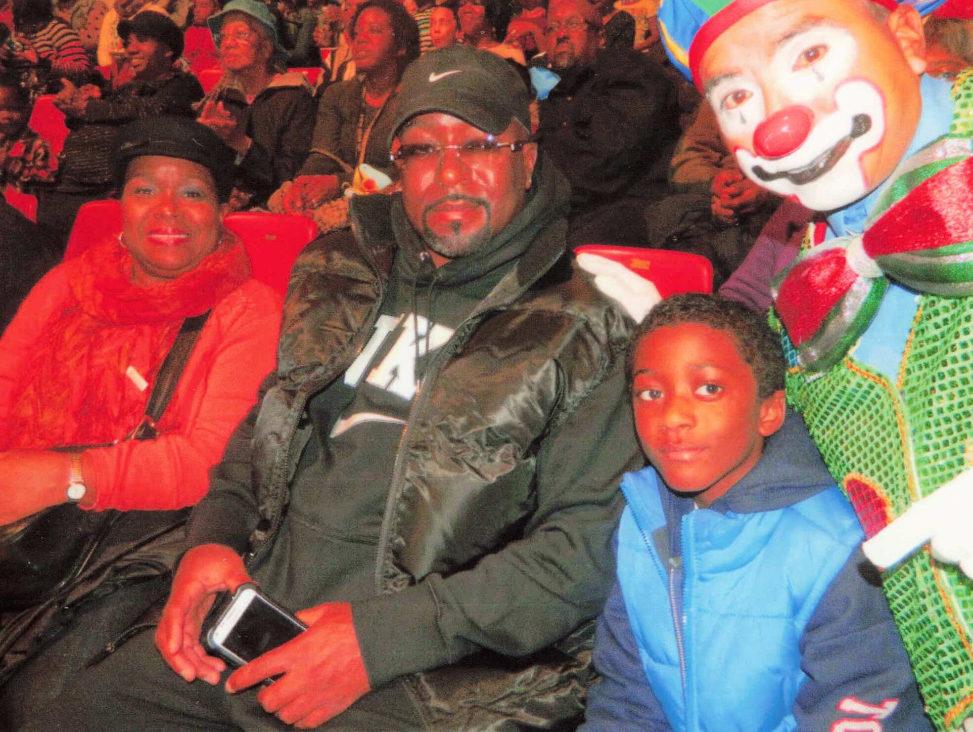 Crowded event scene with a smiling woman in red on the left, a man in a cap and glasses center, a clown in colorful makeup on the right, and a child in a blue jacket in the foreground.