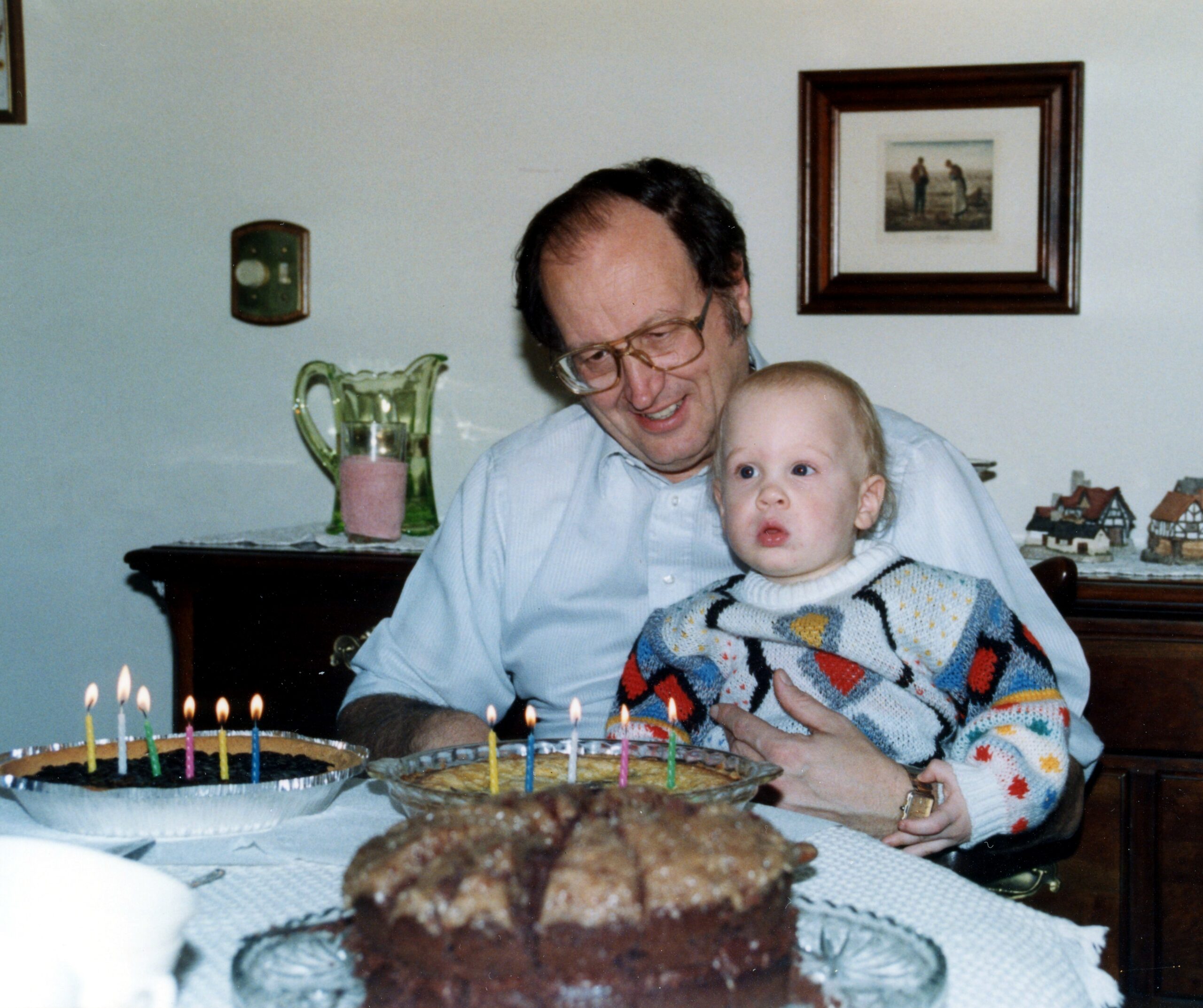 Man smiling while holding a baby at a birthday table with a cake and lit candles in front, indoors at home.