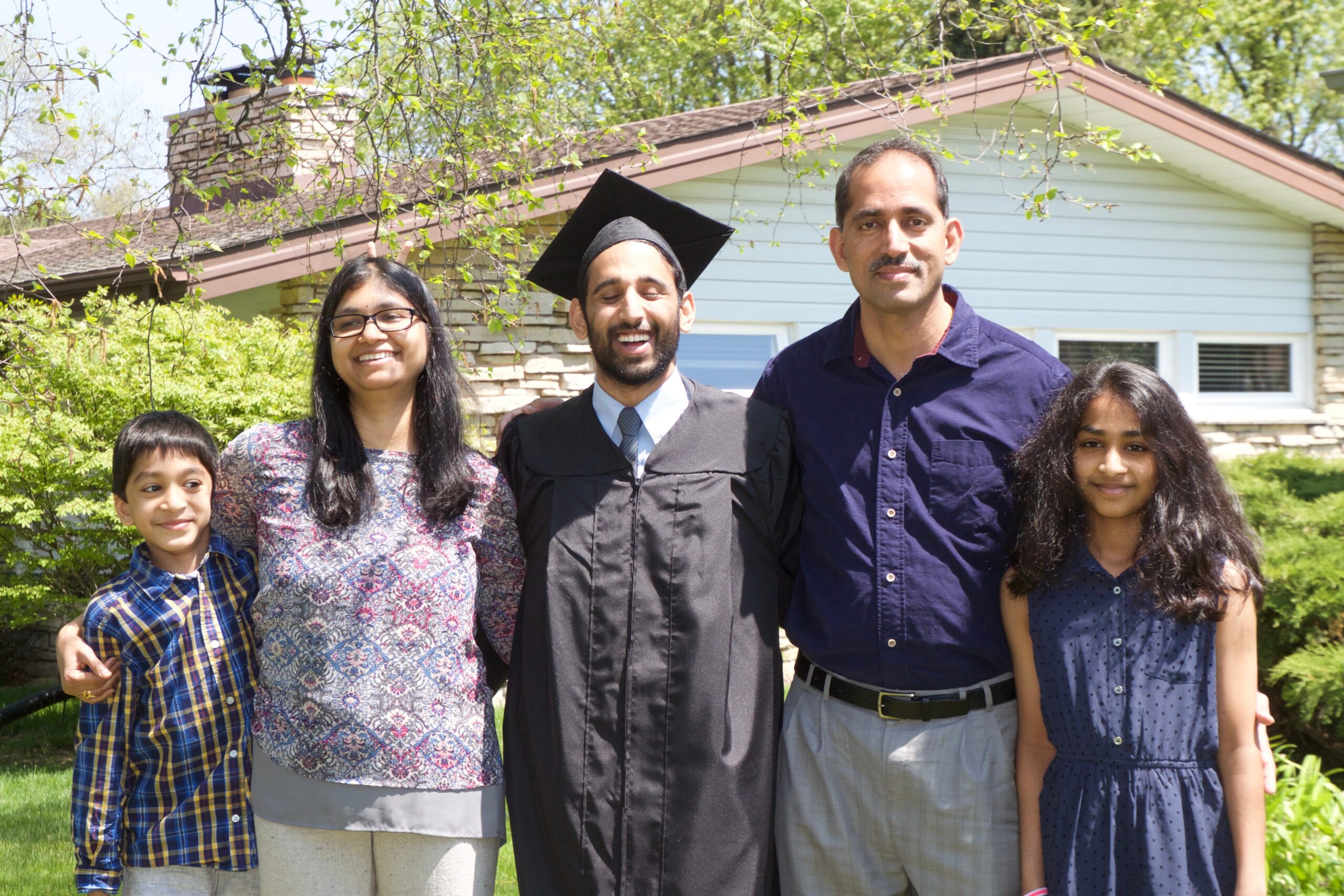 Five people pose outdoors in front of a house and greenery; a graduate in a cap and gown stands in the center with family on either side.