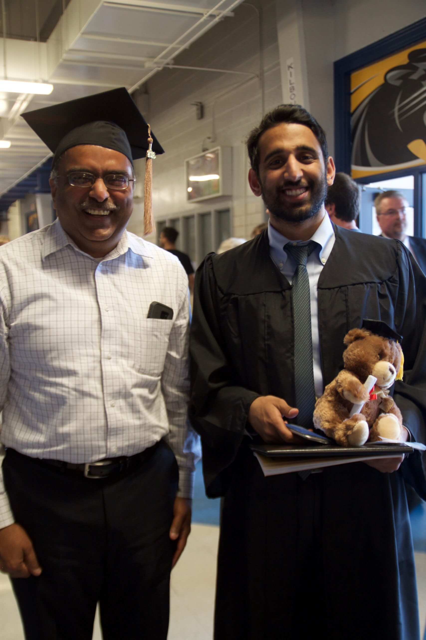 Two men in graduation attire smiling for a photo in a hallway; one wears a cap with tassel, the other holds a teddy bear and folders