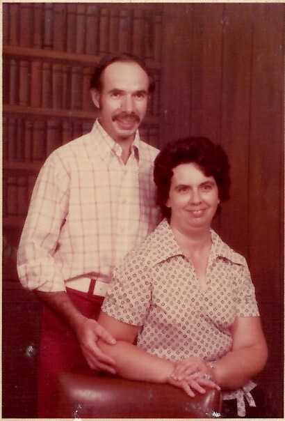 Man with a mustache stands behind a seated woman, both smiling, in a room with a wooden bookshelf.