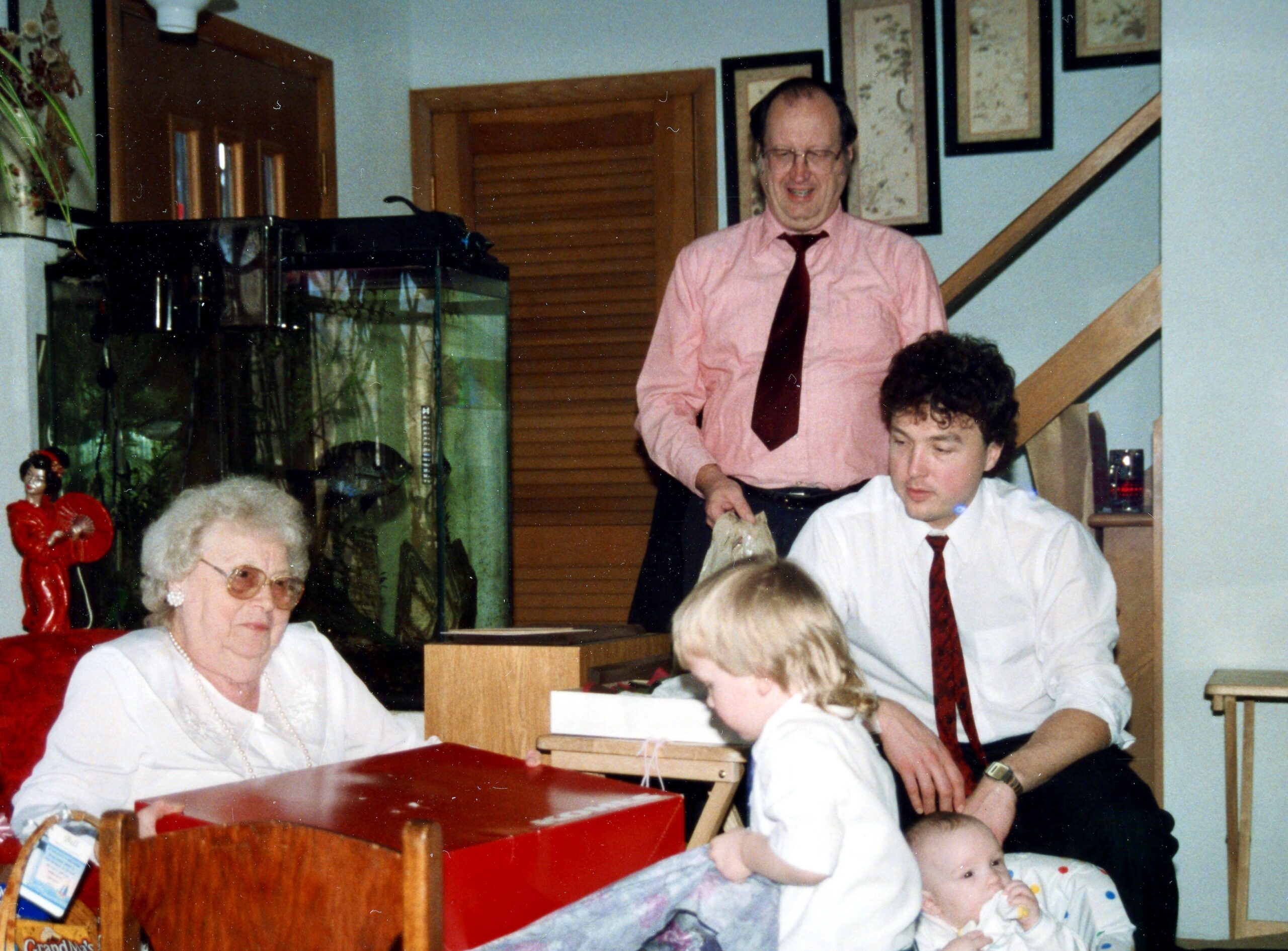 Indoor family gathering: an elderly woman sits at a table while a man in a pink shirt and red tie stands behind, with a baby in the foreground and an aquarium in view behind them.