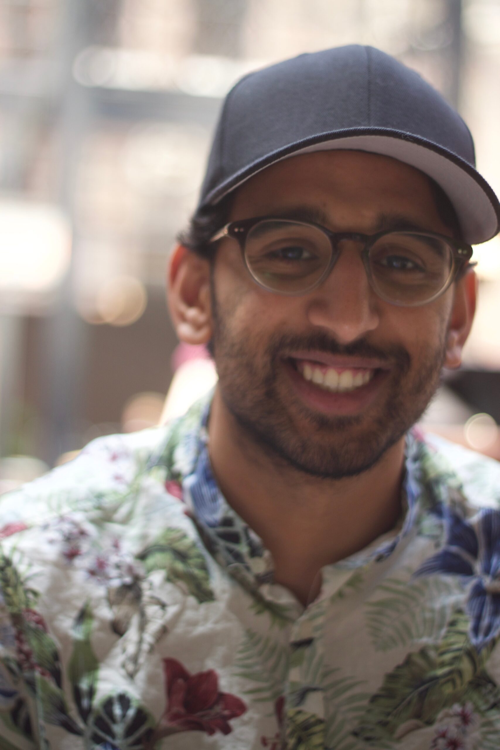 Smiling man wearing a dark cap and glasses, in a floral shirt, outdoors with a blurred background.