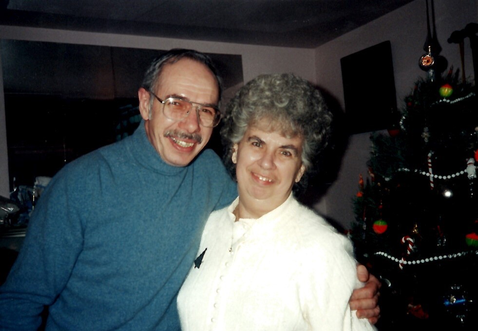 Older couple smiling and hugging indoors, man in a blue turtleneck and glasses, woman in white, with a decorated Christmas tree in the background.