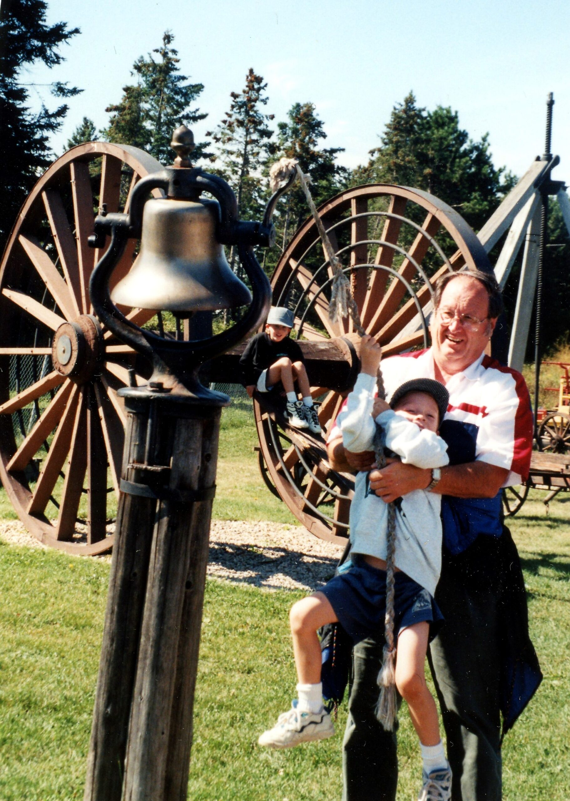Smiling man with two kids swinging on a rope in a park, set against large wooden wagon wheels and a metal bell.