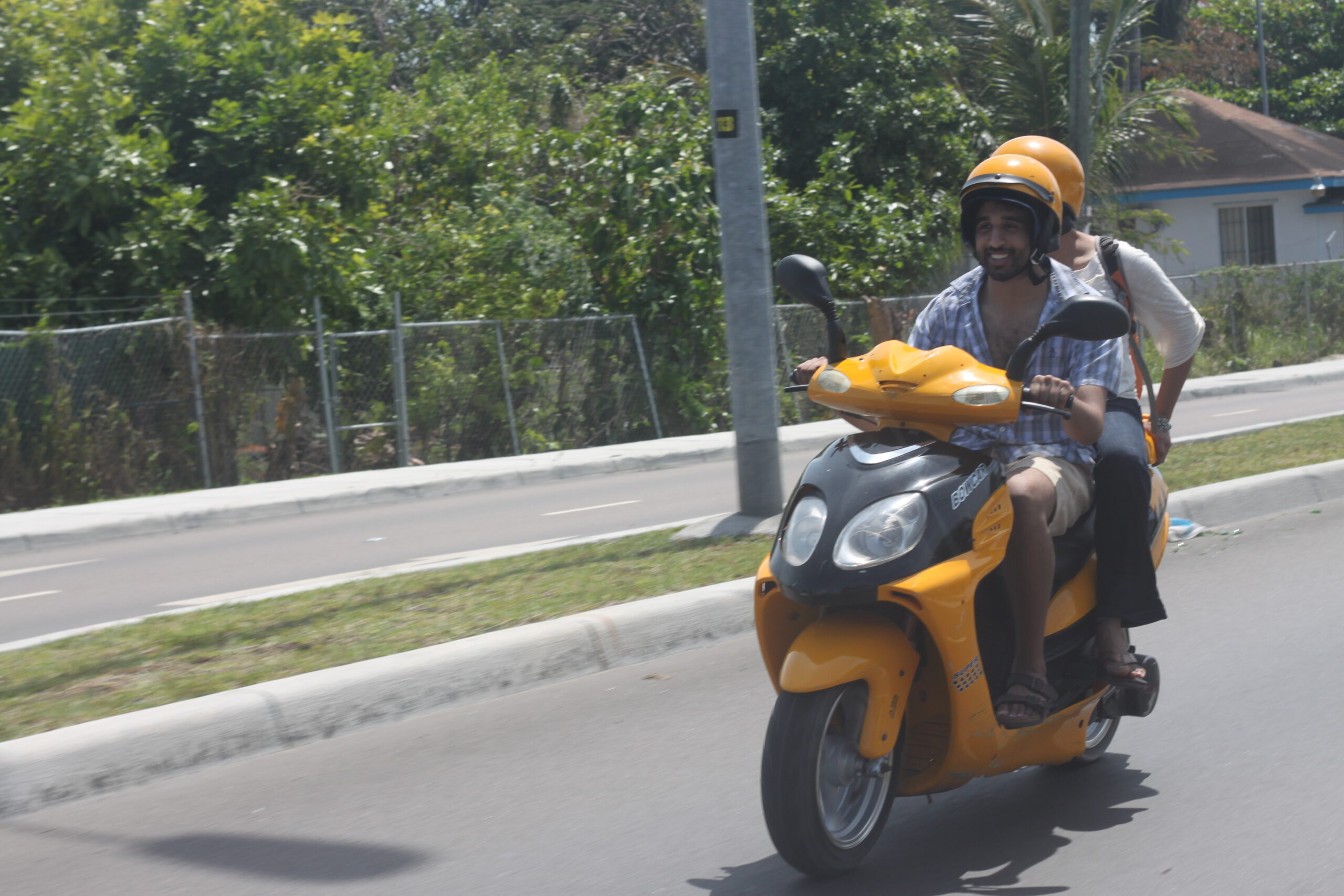 Two people ride a yellow scooter on a sunny road, both wearing helmets and smiling.
