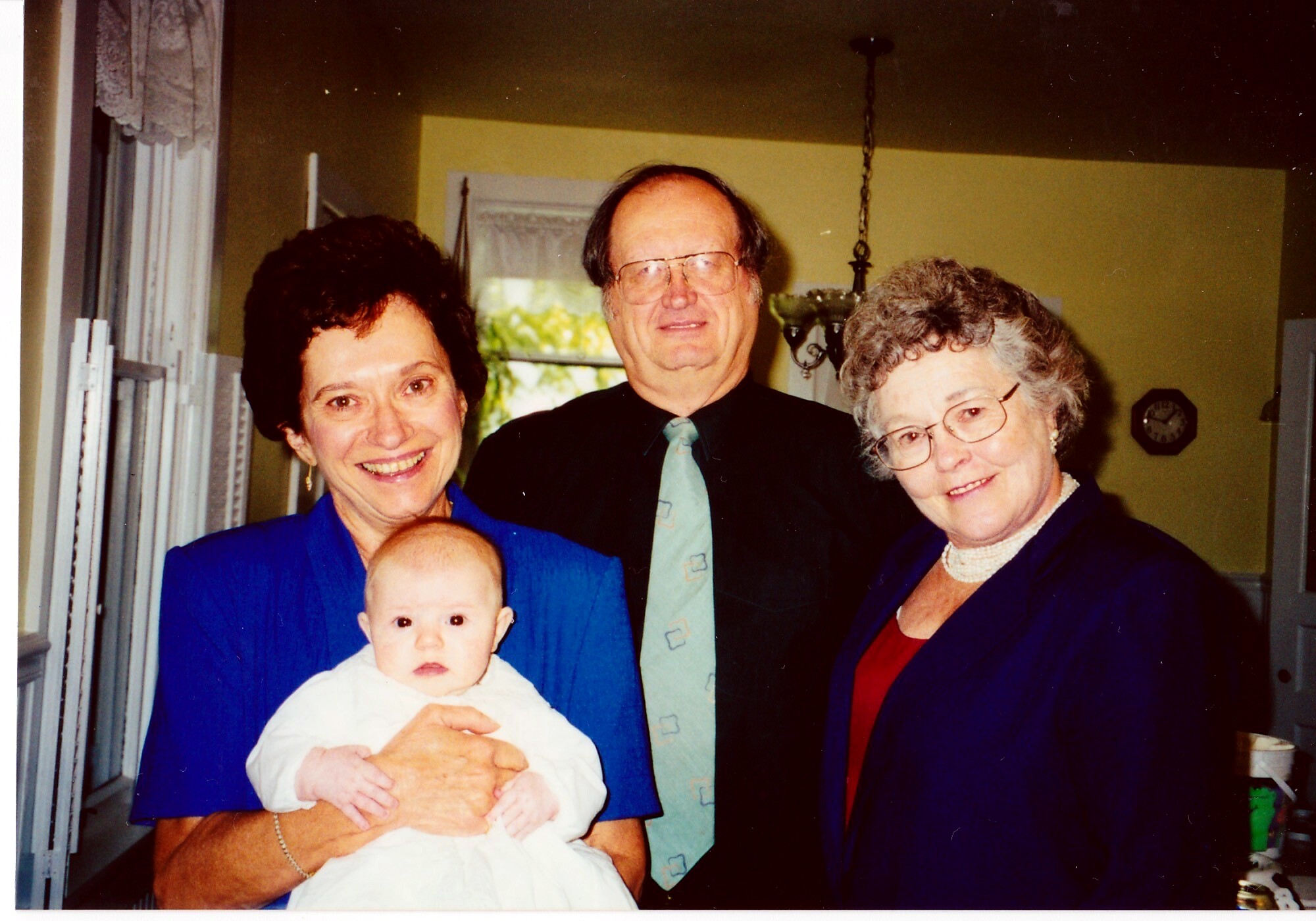 Smiling woman in blue holding a baby, flanked by a man in a suit and an older woman indoors.