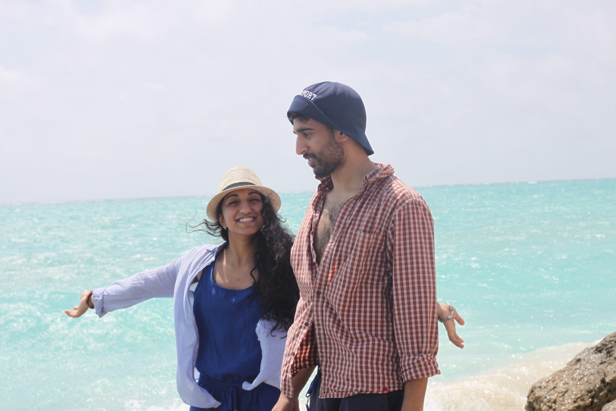 Two friends stand at the beach with arms around each other, smiling, turquoise sea behind them on a sunny day.